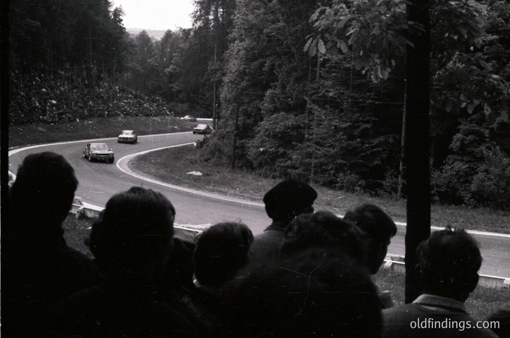 Black-and-white shot of a vintage race car navigating a winding mountain road, surrounded by spectators on the left. Classic racing scene with dense forest backdrop, likely 1960s–1970s.