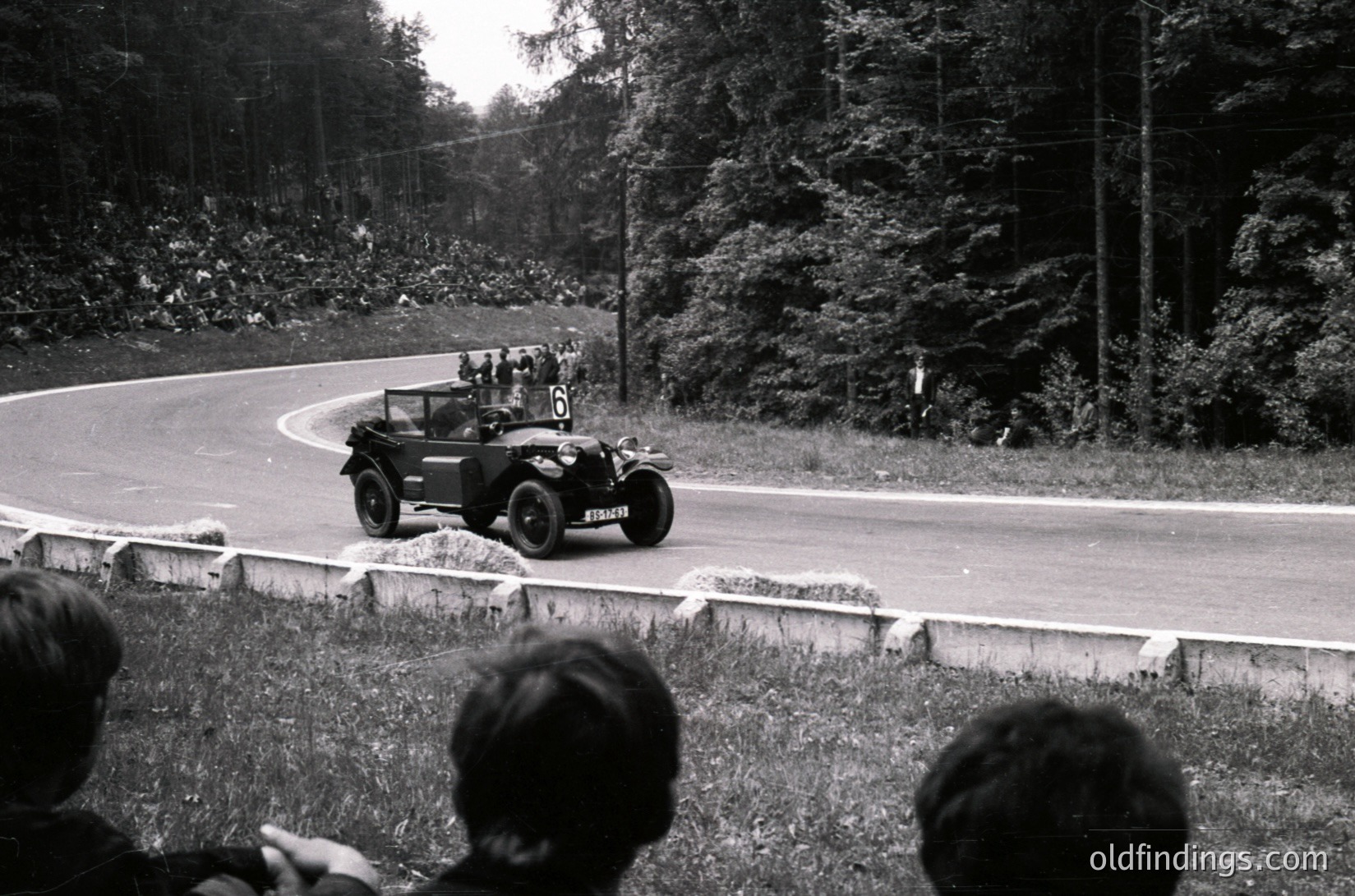 Vintage open-top race car navigating a winding road during a rally, surrounded by spectators. The car’s rounded fenders and streamlined design suggest mid-20th century automotive engineering. Dense forest and grassy embankments frame the scene, indicating a rural or mountainous route.