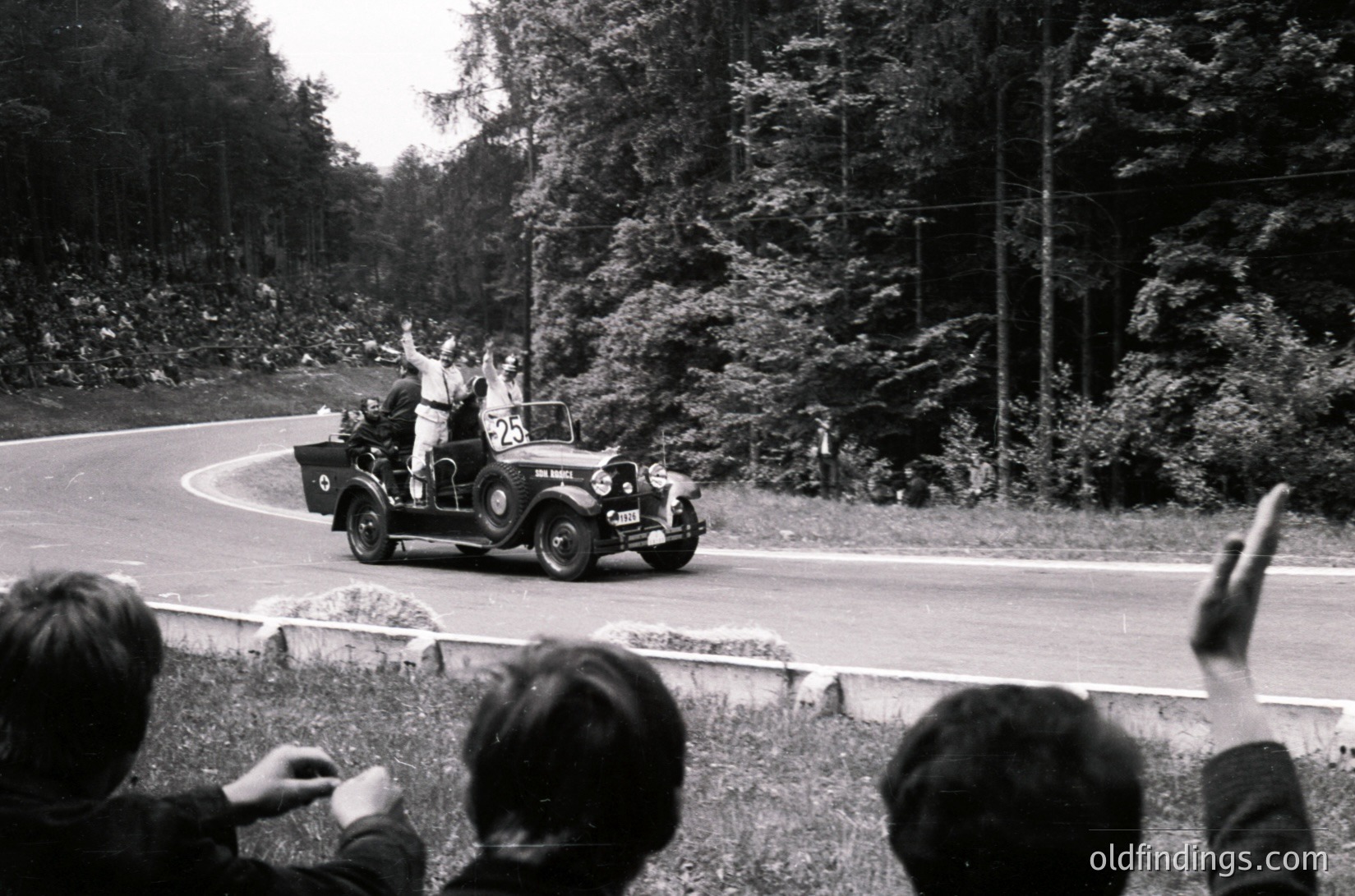 Vintage open-top car navigating a winding forest road, surrounded by onlookers waving. Classic 1930s–1940s roadster with visible license plate and side mirrors. Dense evergreen trees frame the scene, suggesting a rural or mountainous area.
