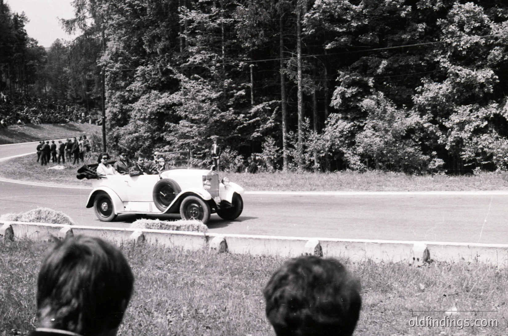 Classic 1930s roadster racing on a rural road, surrounded by spectators. Open-top vintage car with streamlined bodywork and wire-spoke wheels, likely a pre-WWII European model. Lush green forest backdrop and winding asphalt road. Crowd gathered along grassy embankment, indicating public event or rally.
