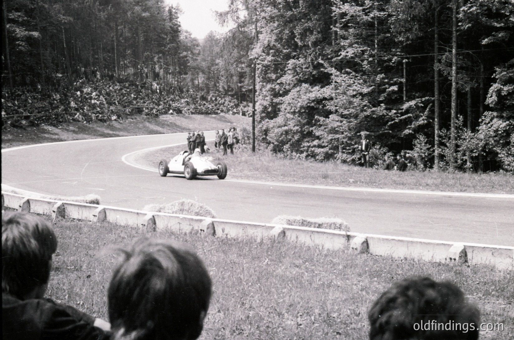Classic open-wheel race car navigating a sharp curve on a wooded road, surrounded by spectators. Mid-20th century racing scene, likely 1950s–1960s. Stone guardrail and dense foliage frame the track.