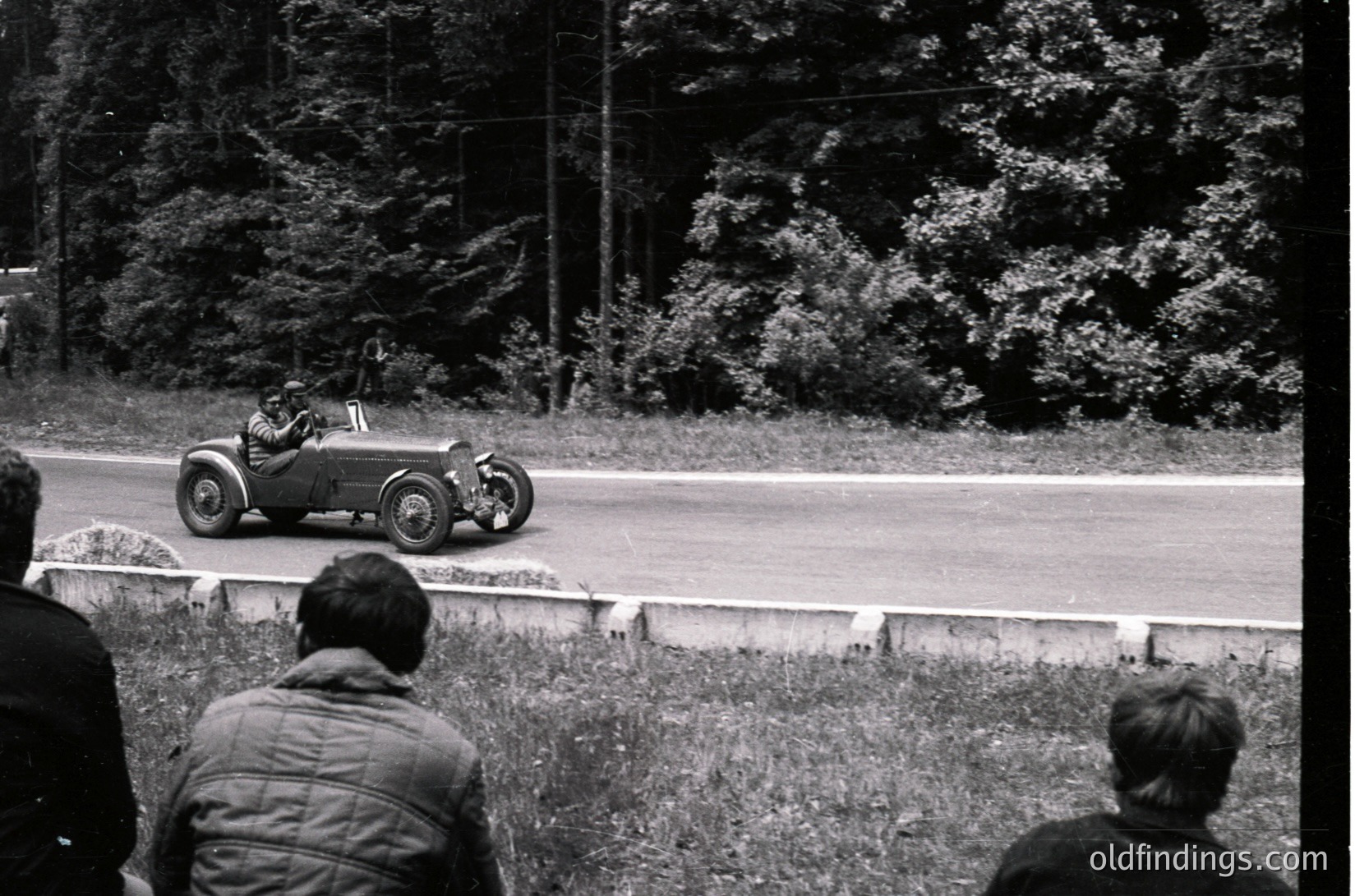 Vintage open-wheel race car navigating a curved road, surrounded by dense forest. Spectators in 1950s-era attire watch from grassy verge. Classic racing scene with mid-century road racing aesthetics.