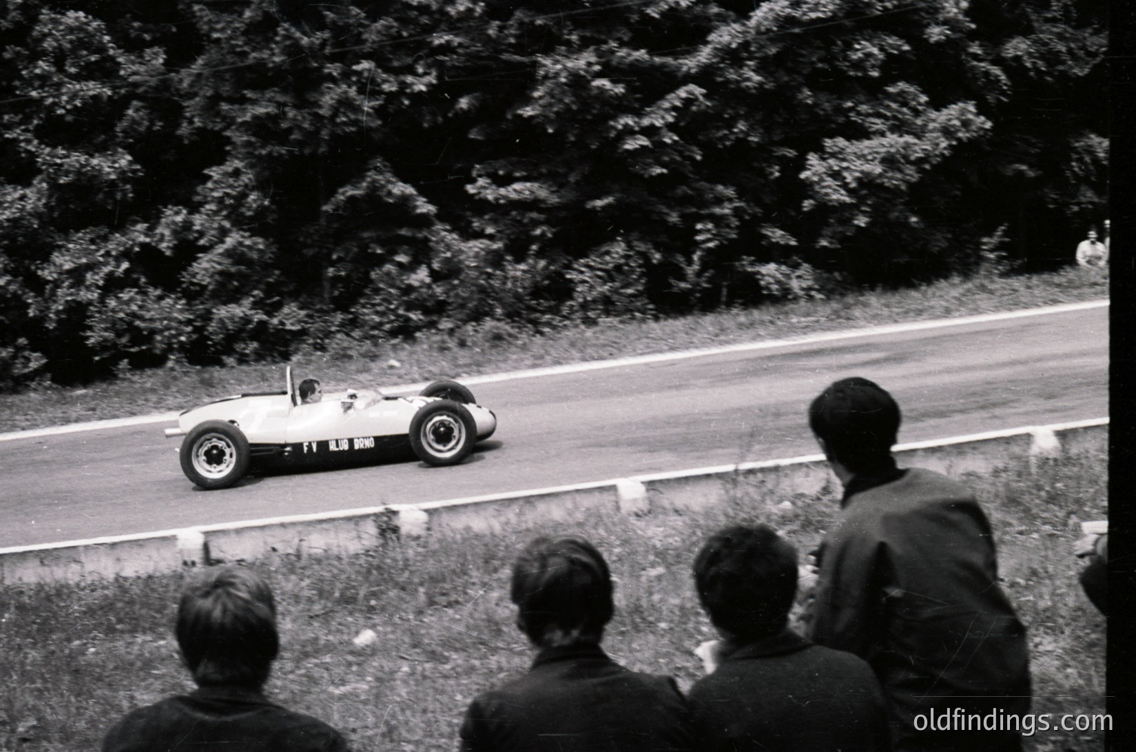 Classic open-wheel race car (Lotus or similar) navigating a winding road, likely during the 1960s. Spectators in dark suits watch from the side. Dense forest backdrop suggests a European circuit. High-contrast black-and-white captures dynamic motion and vintage racing culture.
