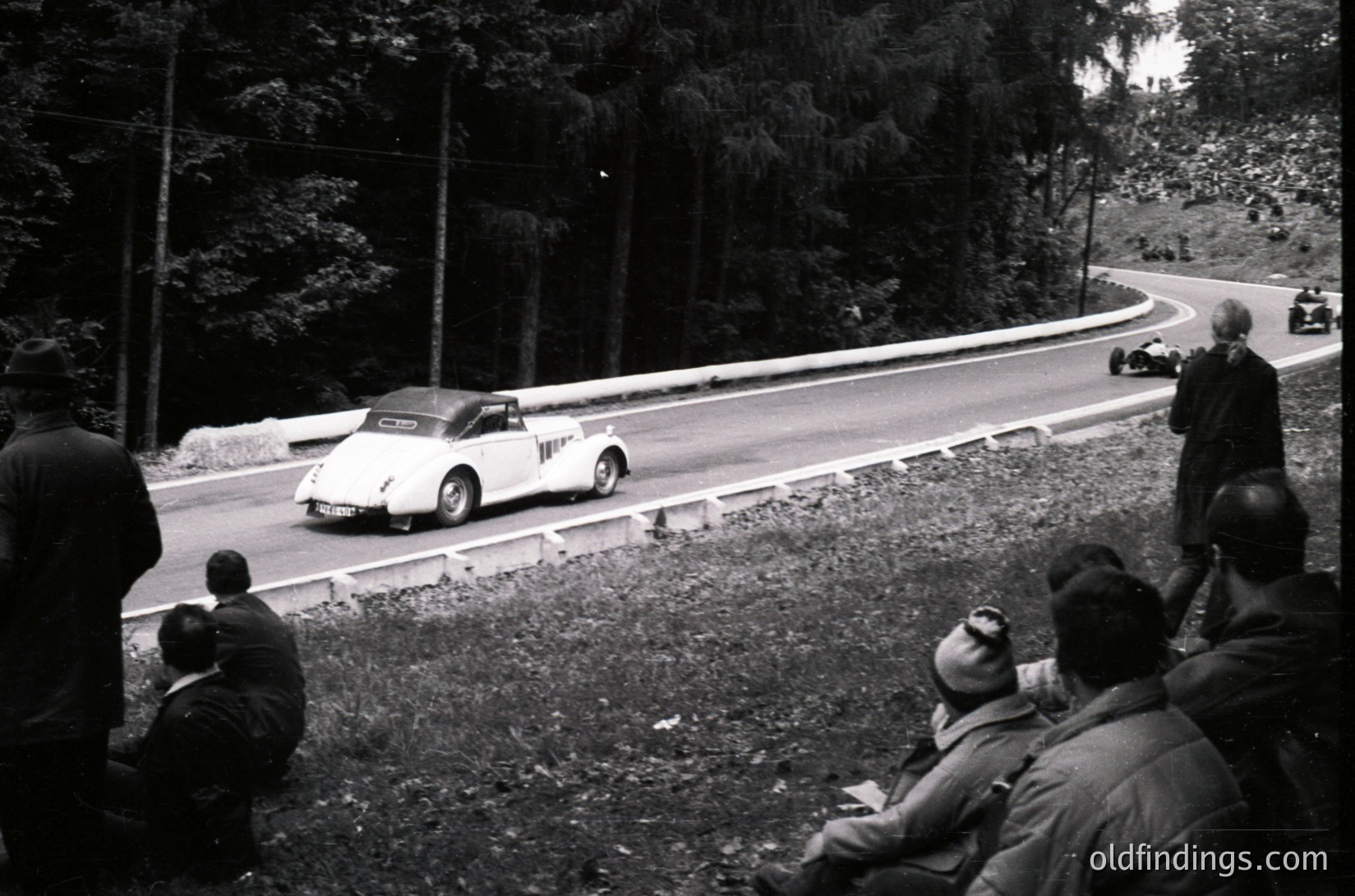 Classic race car navigating a winding forest road during a vintage rally, mid-20th century. Spectators in dark coats line the grassy embankment, watching intently. Dense pine trees frame the scene, emphasizing the road’s curvature.