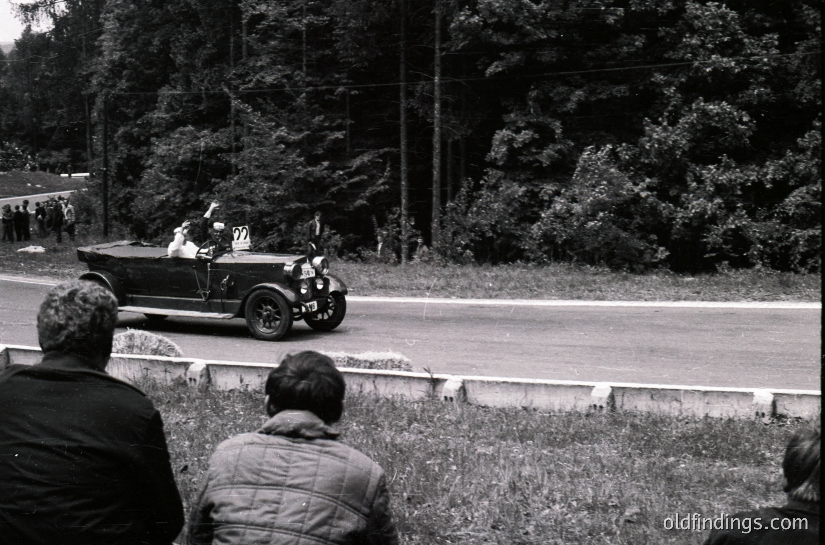 Vintage open-top race car with white racing numbers on black body, likely a classic 1930s-1950s model, driving past spectators on a rural road. Crowd in foreground watches event, surrounded by dense forest. Black-and-white photo captures mid-20th century motorsport culture.