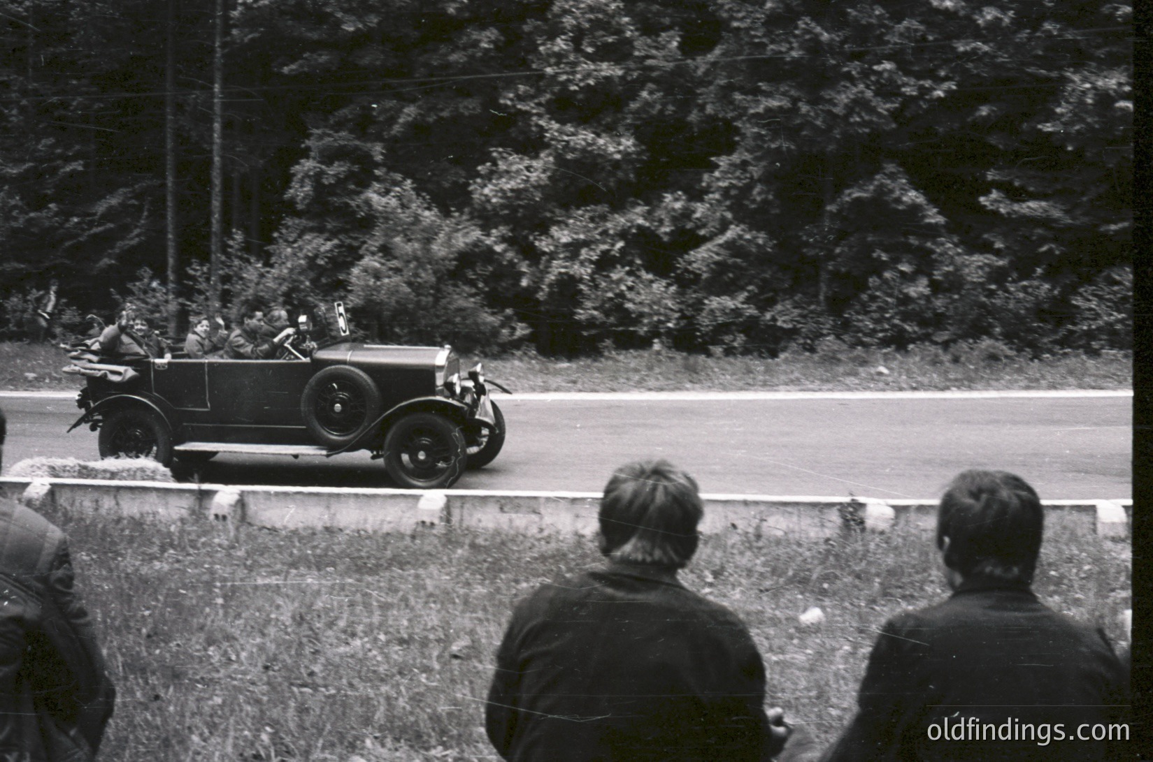 Classic 1930s roadster with open-top design and spoked wheels speeds past spectators on a rural road, framed by dense foliage and a stone wall. Crowd in dark coats watches from grassy verge, suggesting early automotive culture.