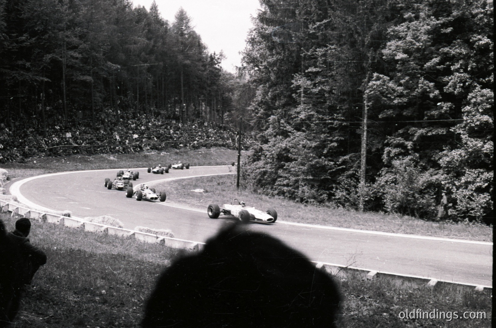 Classic 1960s Formula 1 race at Nürburgring, featuring vintage open-wheel cars navigating a sharp left-hand curve. Spectators in foreground, dense pine forest lining the track. Iconic mid-century motorsport scene. ürburgring