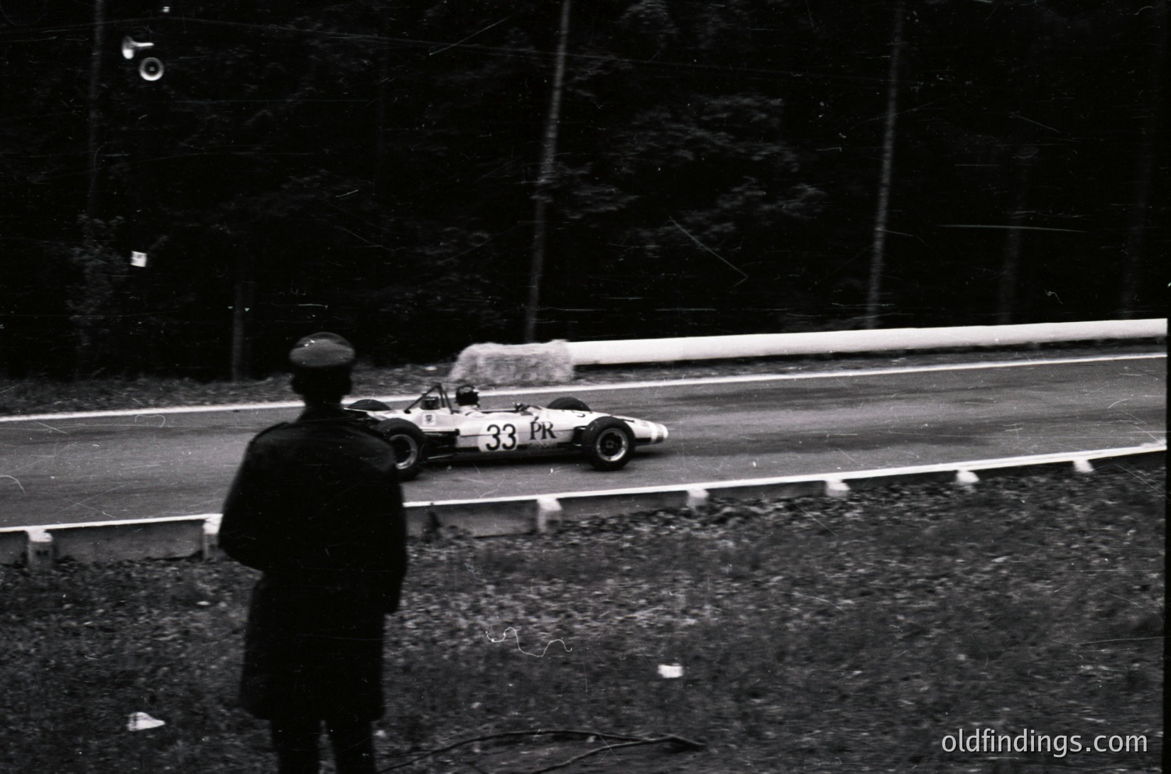 Spectator watches Formula 1 race car () on wet track, likely 1960s–1970s. Open-wheel design, high-speed cornering, and misty conditions suggest classic motorsport era. Ideal for vintage racing archives or automotive history research.