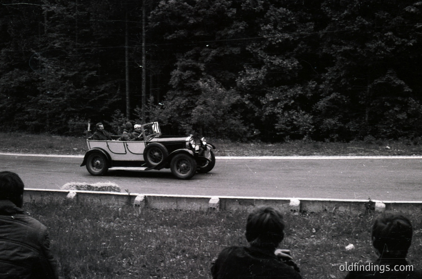 Vintage open-top touring car on a winding road, surrounded by dense forest. Spectators in foreground, likely at a rally or event. Classic 1930s–1940s roadster design with wire-spoke wheels.