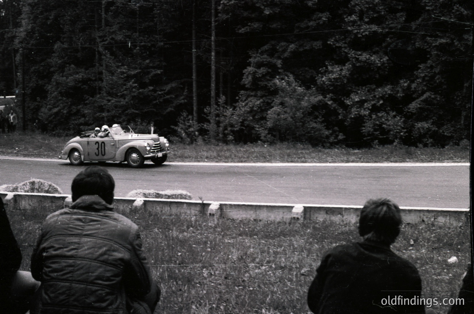 Vintage race car () navigating a winding forest road during a classic rally. Spectators in 1950s-era attire observe from grassy embankment. Black-and-white shot captures mid-century automotive history.