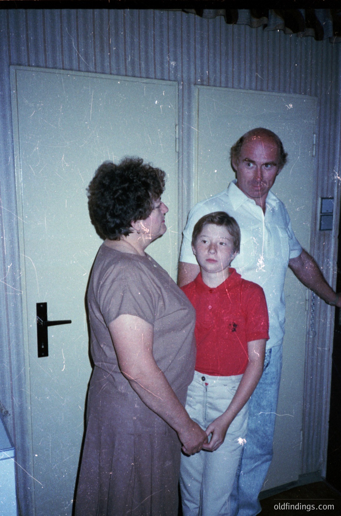 Indoor portrait of three individuals in a dimly lit hallway, likely mid-20th century. Woman in a dark dress stands left, holding a child in a red polo shirt and light jeans. Man in a white shirt and jeans stands right, observing. Faded wallpaper and a door with a black cross symbol in the background.