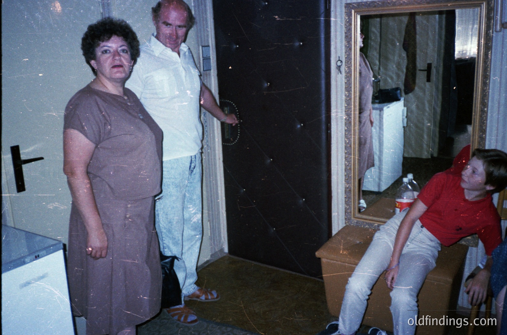 Vintage indoor scene featuring three adults in a dimly lit room with dark paneling. Woman in a brown dress stands beside a man in a white shirt, while a seated man in a red shirt leans against a wooden trunk. Mirror reflects clothing and a can. Mid-20th century domestic setting, likely 1960s–1970s.