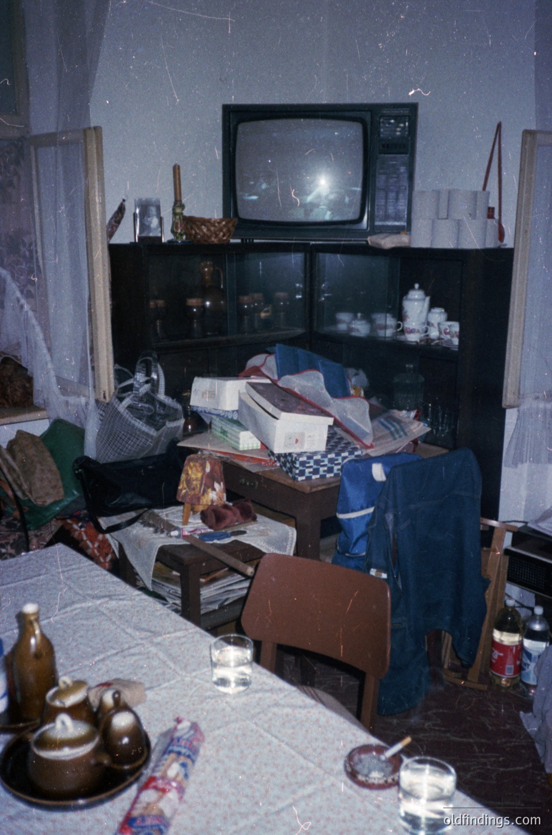 Vintage living room featuring mid-century Soviet-era furnishings: black-and-white checkered tablecloth, wooden cabinet with stacked boxes, and a CRT TV atop a shelf. Decor includes a woven basket, glass bottles, and a blue apron. Cluttered yet nostalgic, indicative of 1970s–1980s domestic life.