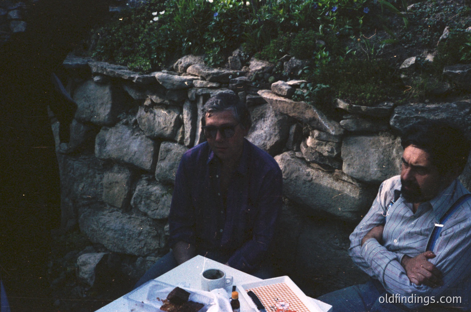 Two men seated outdoors at a stone-walled table, likely mid-20th century. Stone retaining wall with greenery behind them. Table holds a coffee cup, ashtray, and a small notebook. Both wear casual, slightly dated clothing—one in a dark shirt, the other in a light blue button-up.
