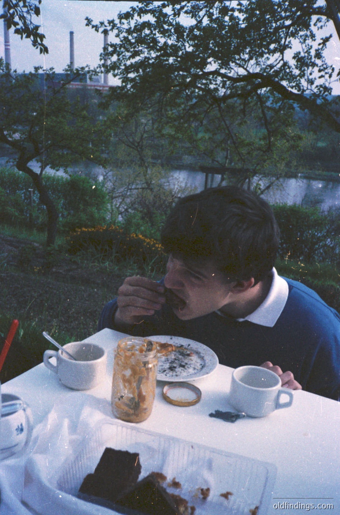 Man eating outdoors by a body of water, mid-1970s. Table set with a half-eaten cake, coffee cup, and disposable cutlery. Industrial chimney and lush greenery in background. Casual, vintage picnic scene.
