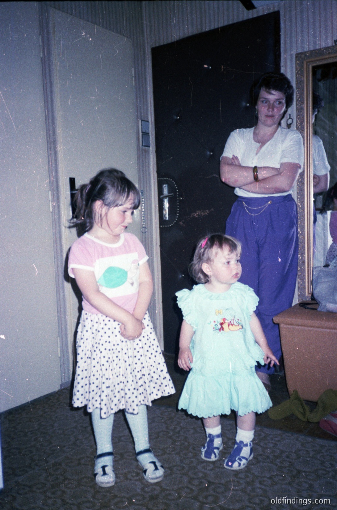 Vintage indoor portrait of two young girls (ages ~4-6) and an adult woman in a modestly furnished room, likely a 1970s-80s home. Girls wear polka-dot dresses with matching sandals; one has a circular patch on her blouse. Woman stands arms crossed, wearing a white blouse and patterned skirt. Dark door frame and worn carpet suggest utilitarian decor.