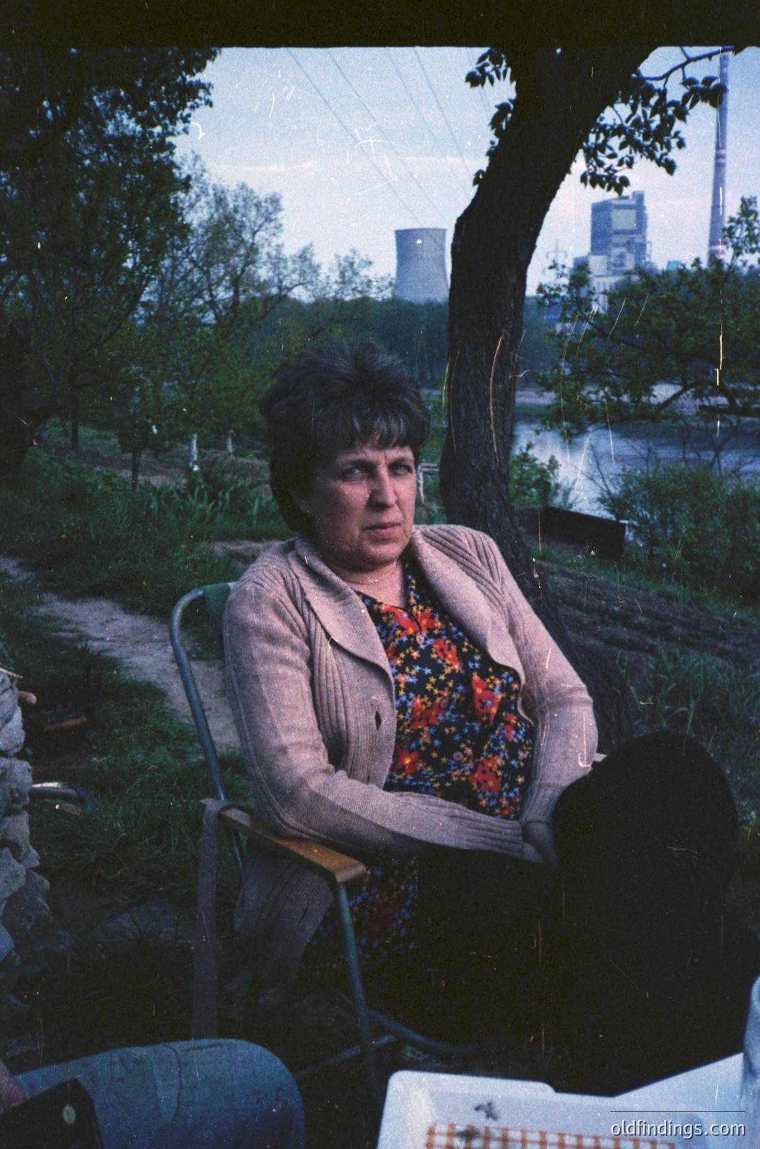 Woman seated outdoors on a metal chair, wearing a patterned blouse and beige jacket, with industrial buildings and greenery in the background. Likely late 20th century, urban/suburban setting.