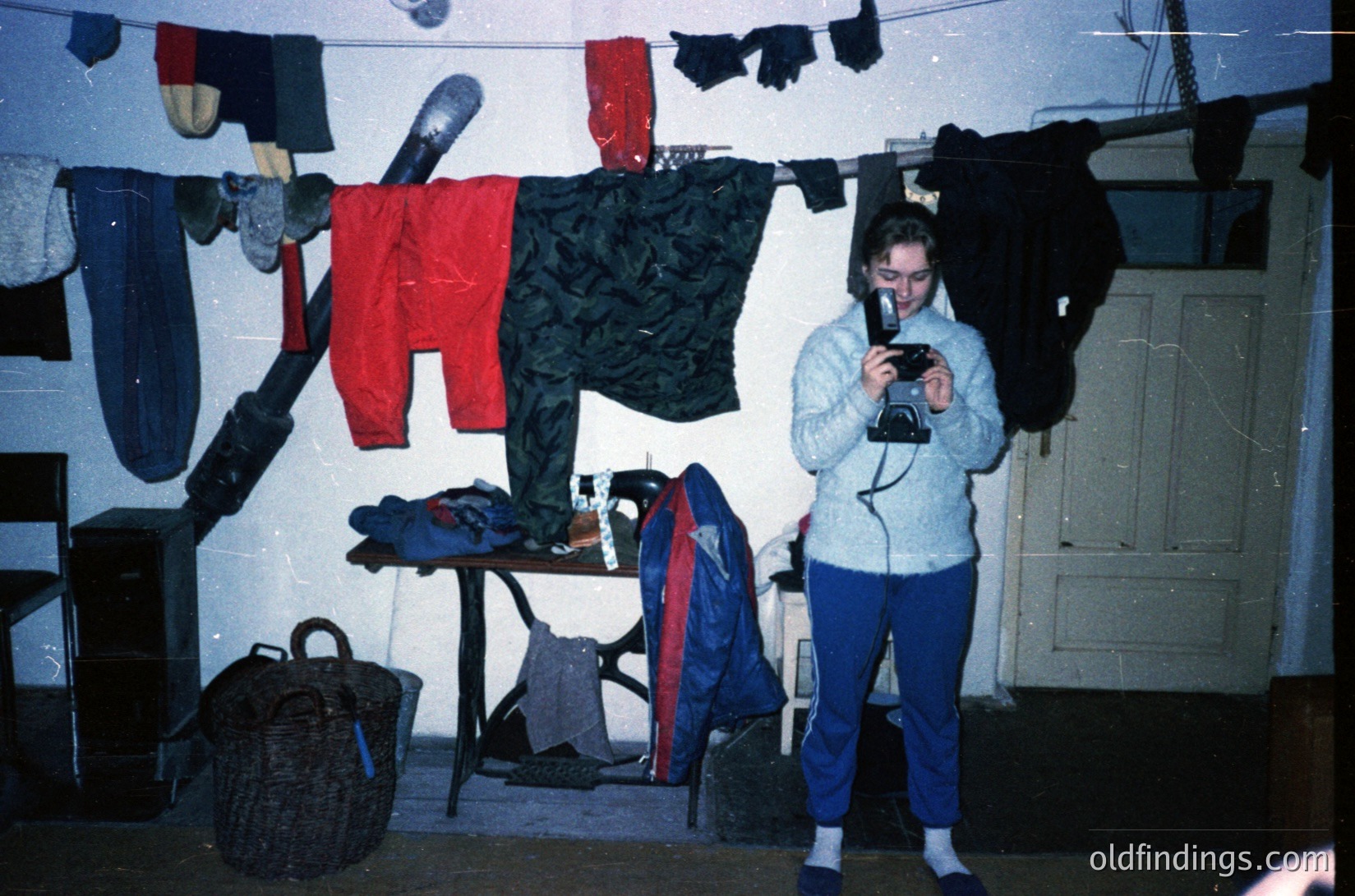 Vintage indoor laundry scene with 1970s-era clothing hung on lines. Person in a light sweater and blue pants holds a bulky camera, likely a Polaroid. Items include red pants, black knitwear, and a wicker basket. Bright overhead lighting highlights textures and colors.
