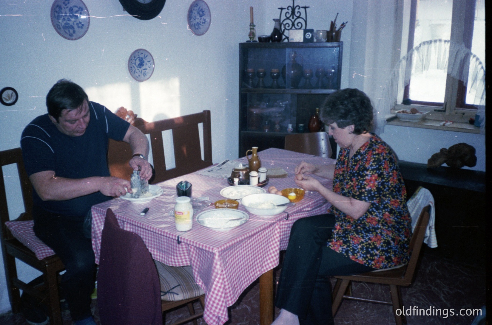 Two individuals seated at a vintage wooden table in a mid-century home, set with checkered tablecloths and ceramic dishes. The man in a striped polo shirt and the woman in a floral blouse engage in conversation. Decor includes blue-and-white wall plates, a glass-front cabinet with decorative items, and a window sill adorned with a woven basket. Likely Eastern European domestic setting, 1960s–1970s.