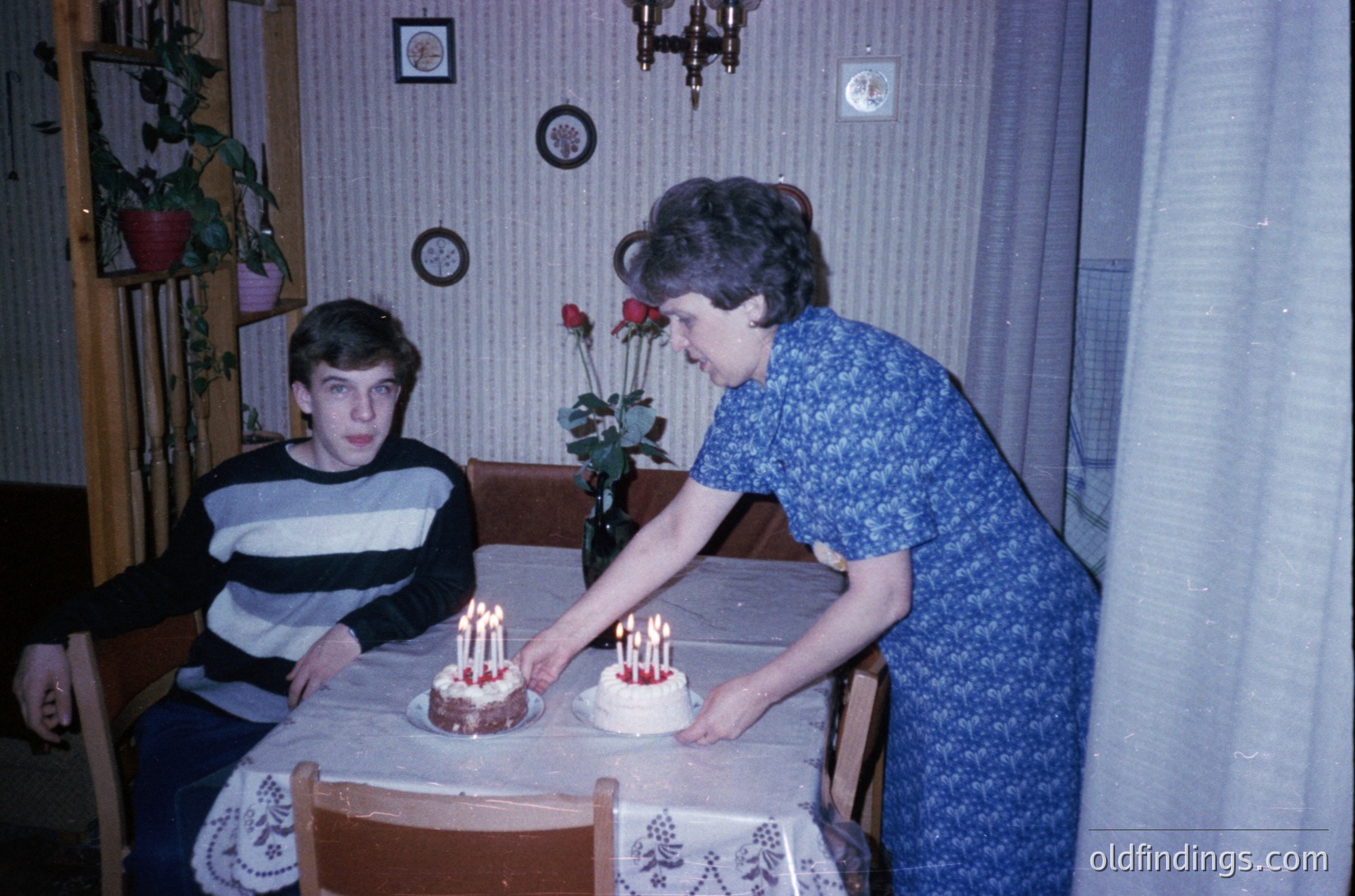 Vintage indoor birthday scene: woman in patterned blouse placing candles on two tiered cakes on a wooden table; teen boy seated beside floral tablecloth. Decor includes potted plants, wall clocks, and striped wallpaper. Likely late 20th century domestic setting.