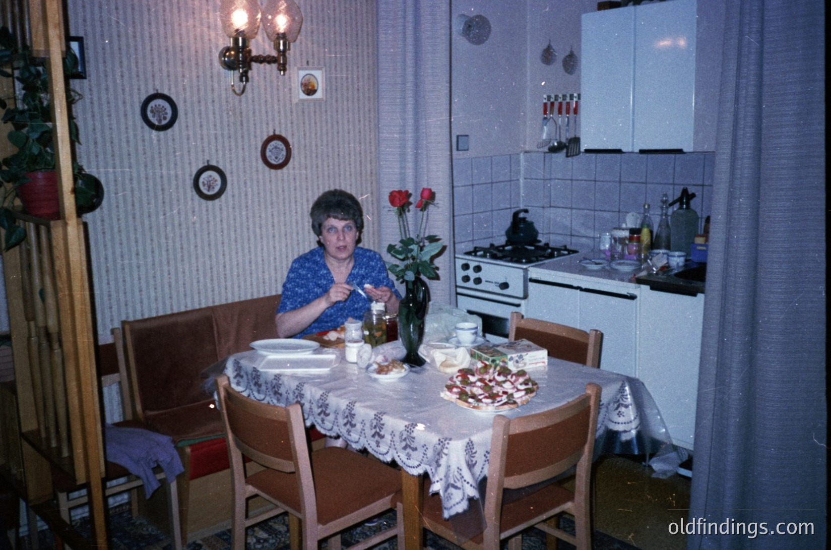Vintage kitchen scene featuring a woman seated at a round wooden table draped with a lace tablecloth, holding a glass. Table set with plates, a vase of red roses, and a bowl of food. White-tiled walls, retro stove, and wall-mounted light fixtures. Mid-century European home decor, likely 1960s–1980s.