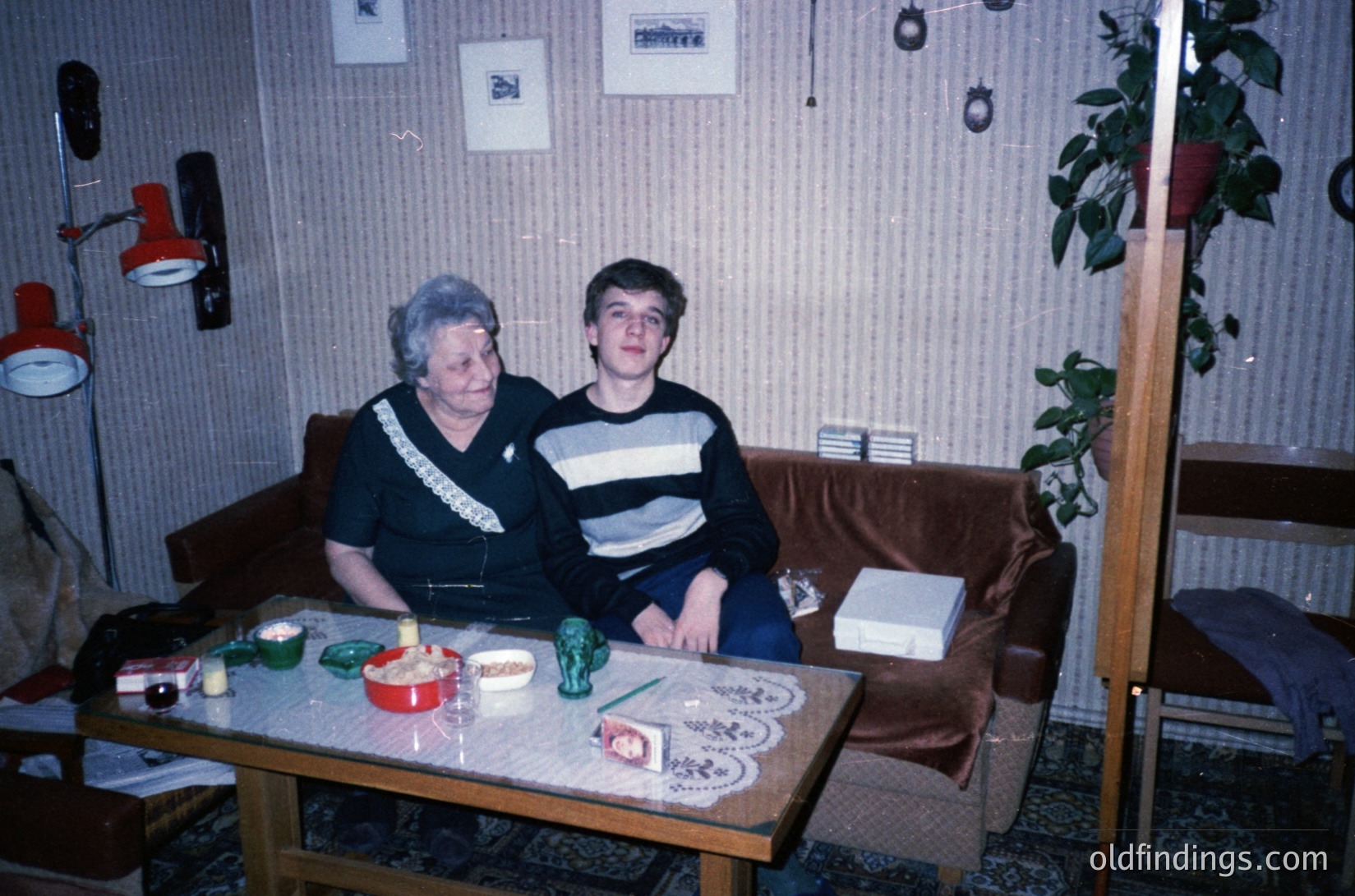 Vintage indoor portrait of an elderly woman and young man seated at a small wooden table in a modestly furnished room, likely Eastern Europe, 1970s–1980s. Tabletop features embroidered textiles, a green figurine, and a framed photo. Wall-mounted lamps and patterned wallpaper suggest utilitarian design. Warm lighting and candid expressions evoke nostalgia.