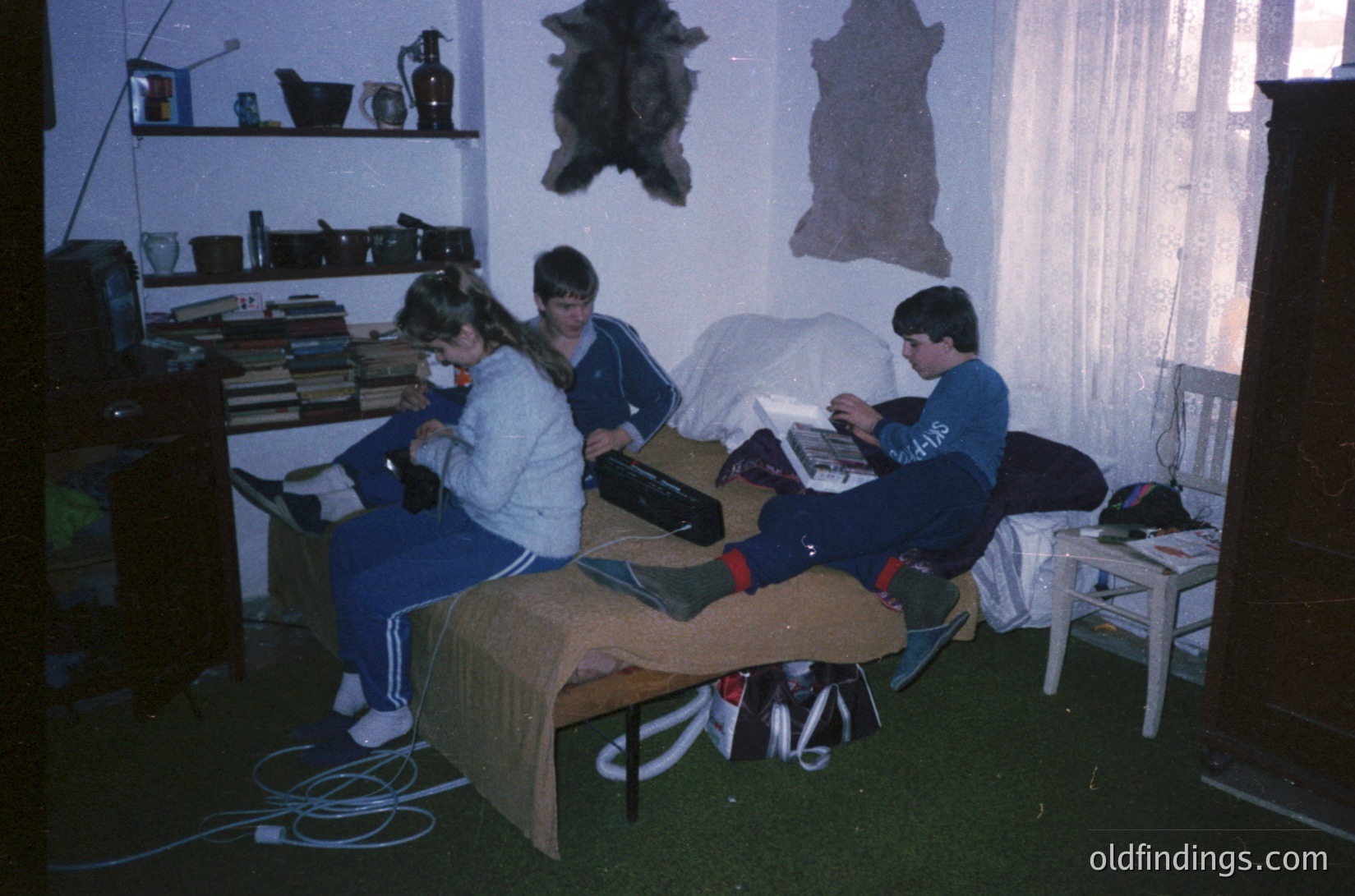 Three individuals study or work at a makeshift desk in a dimly lit room, likely a dormitory or shared living space. The table is covered with books, papers, and a vintage laptop. Animal pelts hang on the wall, and shelves hold various items. The setting suggests a 1990s-early 2000s timeframe, possibly Eastern Europe. [Vintage dorm study session with 1990s-era laptop, animal pelts on wall, and bookshelves. ]