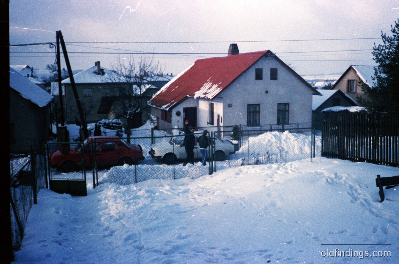 Snow-covered residential street with mid-century concrete houses, red-tiled roofs, and chain-link fences. Two individuals in winter coats near a parked car. Overhead power lines and bare trees suggest late winter or early spring. Likely Eastern European setting.