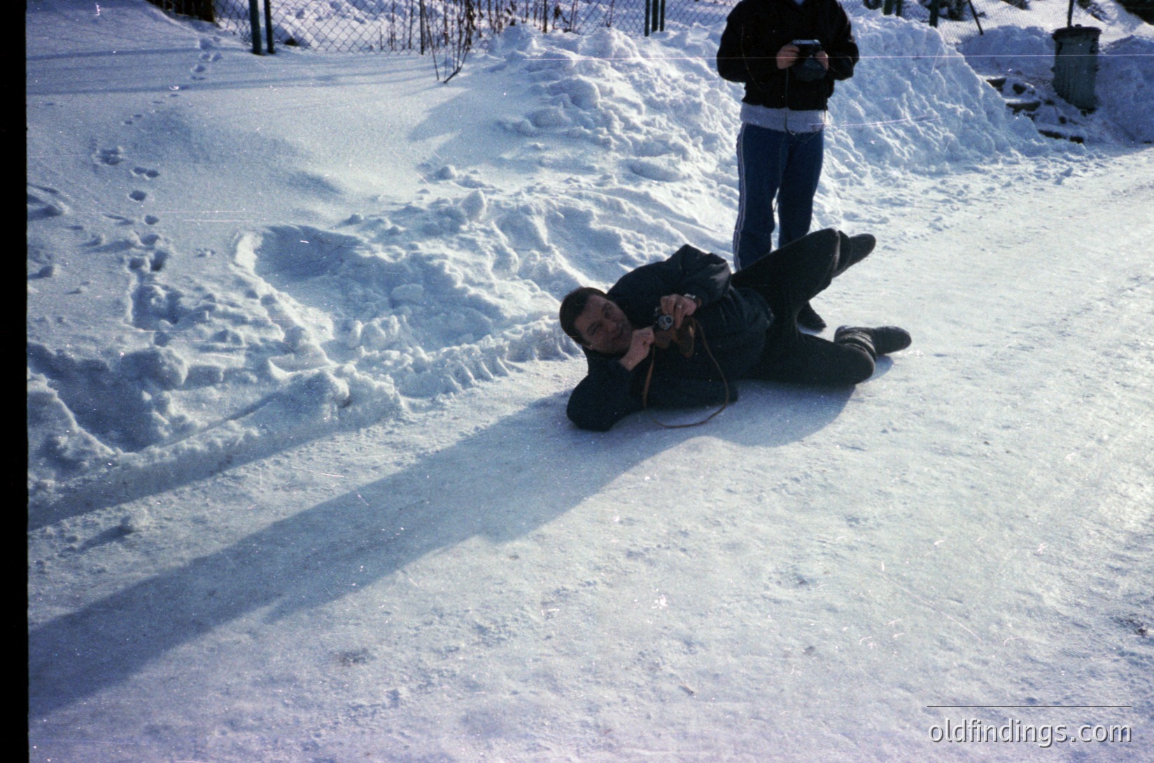 Vintage photo of two men in a snow-covered yard, likely mid-20th century. One man kneels in snow, holding a camera; the other stands, holding a camera and wearing a white scarf. Fence and shed in background suggest residential setting.