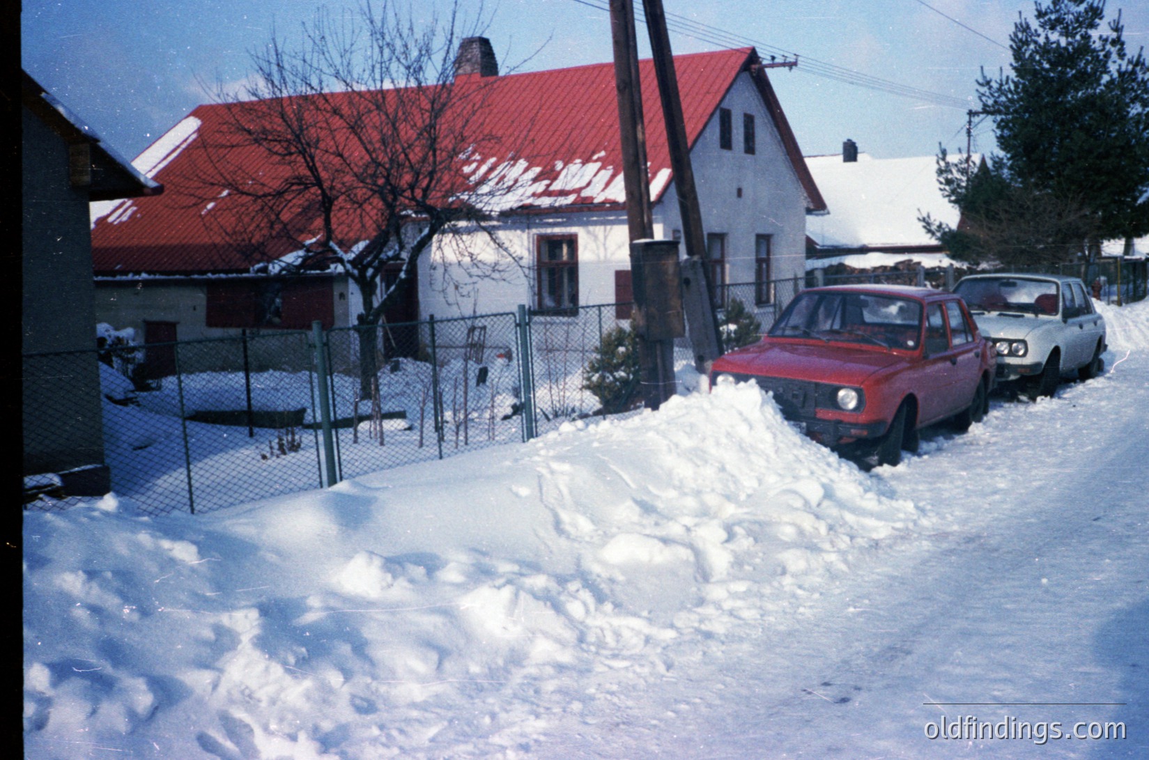 Snow-covered residential street with 1970s-era cars, likely Eastern Bloc region. Whitewashed houses with red-tiled roofs and chain-link fences. Classic compact cars parked on snow-packed driveway.