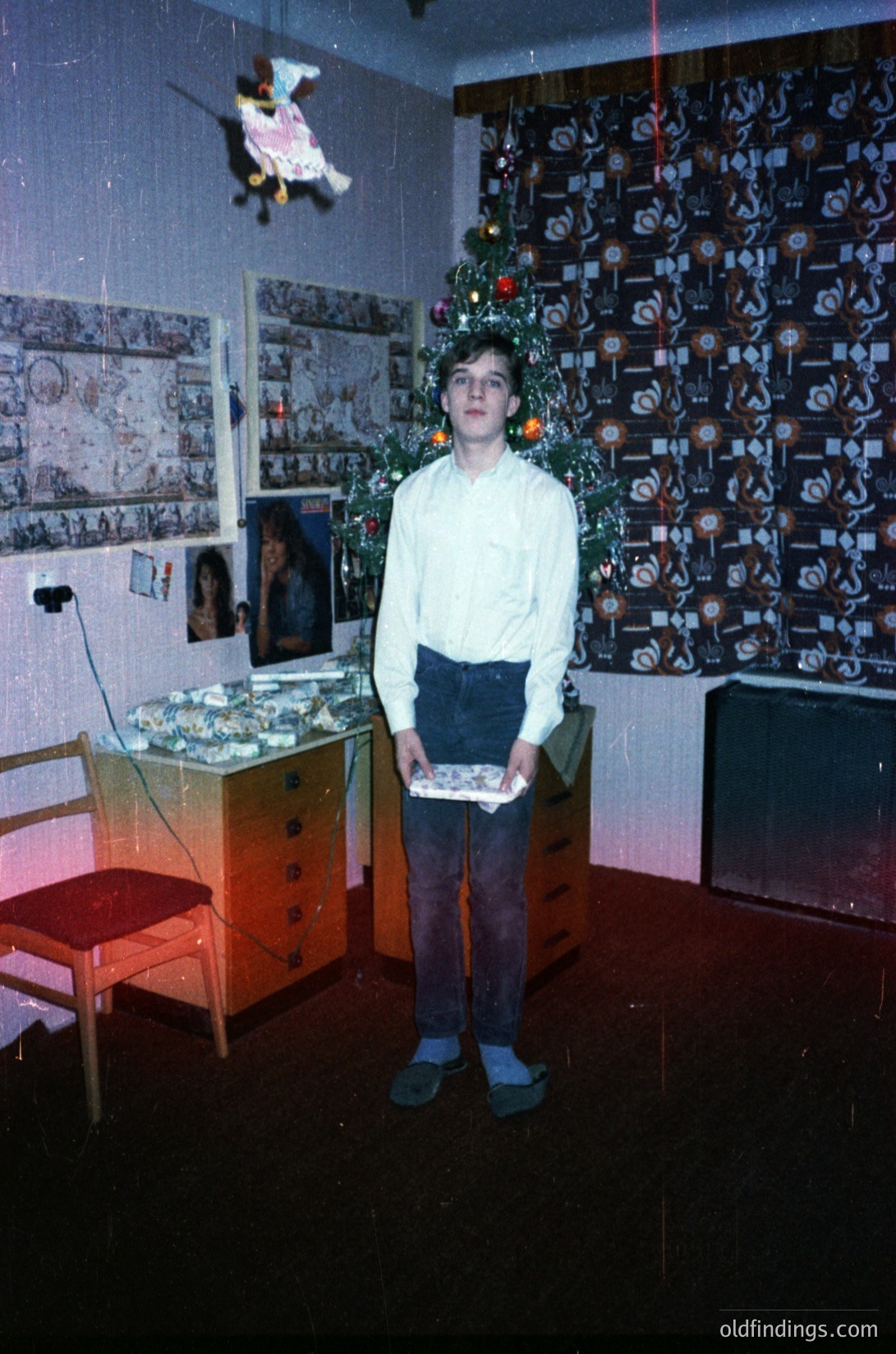 Young boy in 1970s-style sweater and rolled pants poses indoors, holding a wrapped gift. Surroundings include a vintage wooden dresser, a Christmas tree with ornaments, and a wall adorned with framed photos and a decorative paper collage. Warm holiday lighting enhances the nostalgic atmosphere.
