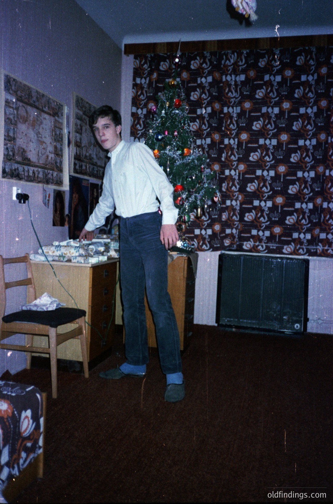 Young man in 1970s-style dress shirt and dark trousers poses in a modestly decorated living room. A vintage Christmas tree adorned with paper ornaments and a single bulb lights the corner. Behind him, a wall covered in framed photos and posters. Side table holds a radio or small stereo, and a black-and-white TV sits on a wooden stand. Warm, nostalgic 1970s home ambiance.