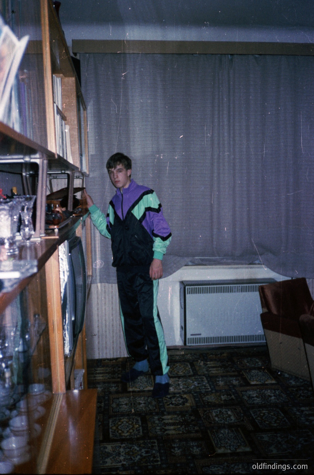 Vintage kitchen scene featuring a person in 1970s-style overalls (purple/blue/green) organizing glassware on a wooden shelf. White electric heater and tiled floor with floral pattern. Warm, nostalgic lighting suggests domestic life.