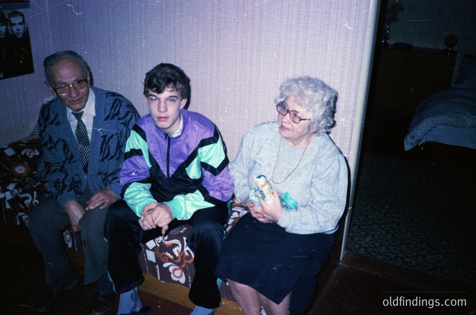 Three generations seated indoors, likely late 20th century. Elderly man in patterned sweater and tie, middle-aged woman in glasses holding a small bird, and young man in neon track jacket. Dim lighting, floral-patterned wallpaper, and a visible bed in background suggest a modest home setting.