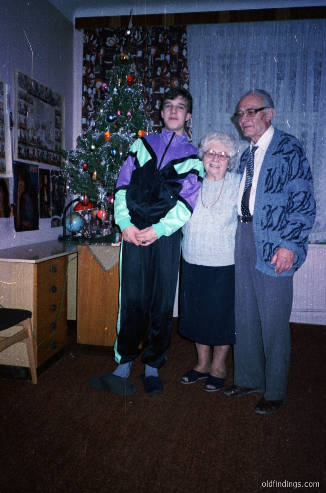 Three generations pose indoors beside a decorated Christmas tree, 1970s–1980s. Teenager in purple sweater and black pants holds a black bag; elderly man in patterned suit and tie stands beside a woman in a white sweater. Wooden dresser, framed photos, and vintage decor suggest a mid-century home. Warm holiday lighting enhances nostalgic family atmosphere.