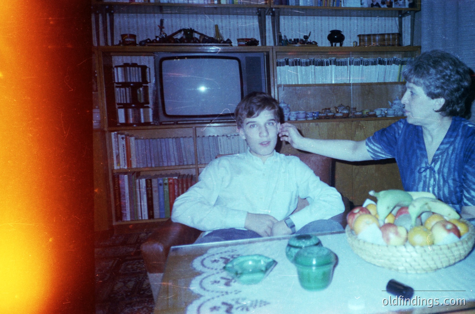 Vintage indoor scene featuring a young boy and adult woman in a mid-century living room. Wooden bookshelves filled with vinyl records and books flank a CRT TV. A table displays a woven fruit basket, vintage glassware, and a small radio. Warm lighting and retro color tones suggest 1960s–1970s domestic life.