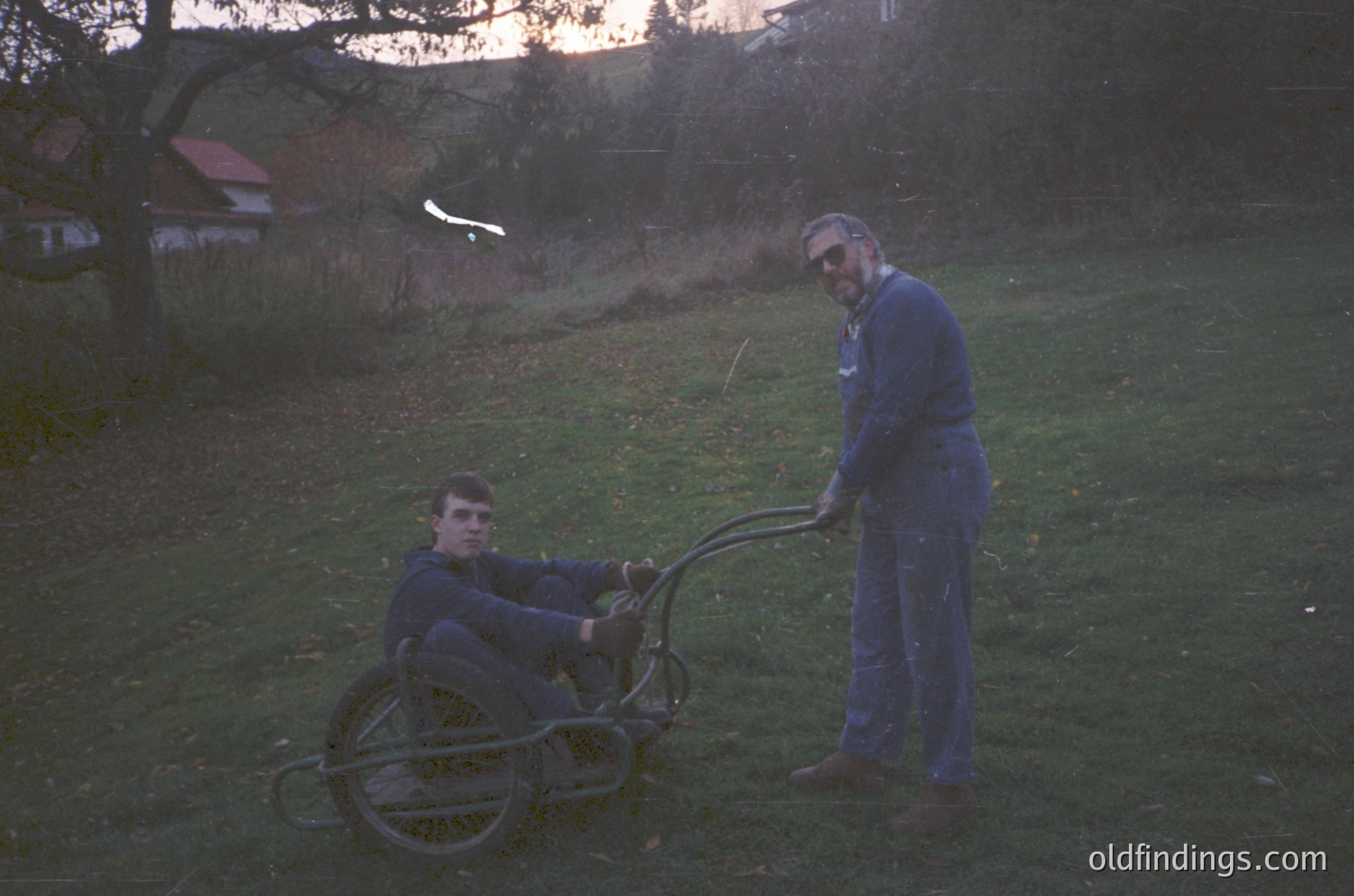 Two men in matching blue workwear assist a manual wheelchair in a rural setting, likely late 20th century. The standing man holds the wheelchair frame, while the seated man appears to adjust the footrests. Low-angle lighting suggests early morning or late afternoon. The background features sparse trees and a distant house, evoking a pastoral or agricultural context.