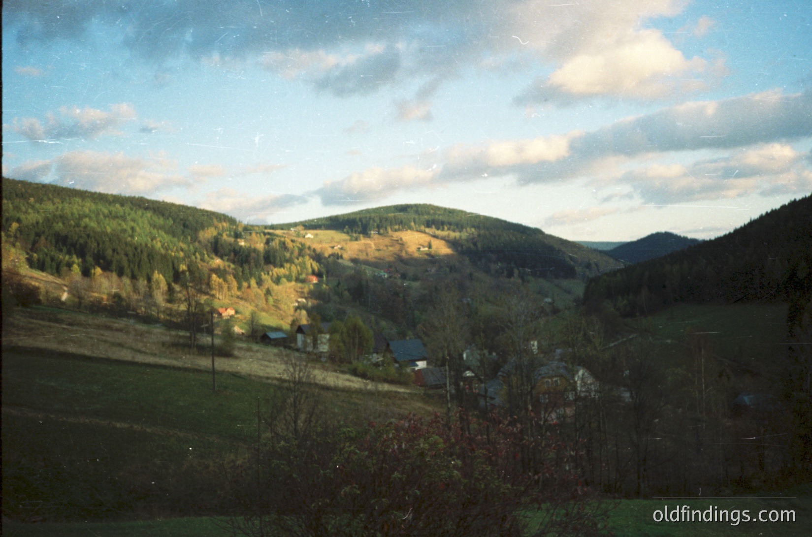 Vintage landscape shot of rolling hills with autumn foliage, sparse farmhouses, and dense forest. Soft color tones suggest mid-20th century film. Rural European countryside, likely Eastern Bloc region.