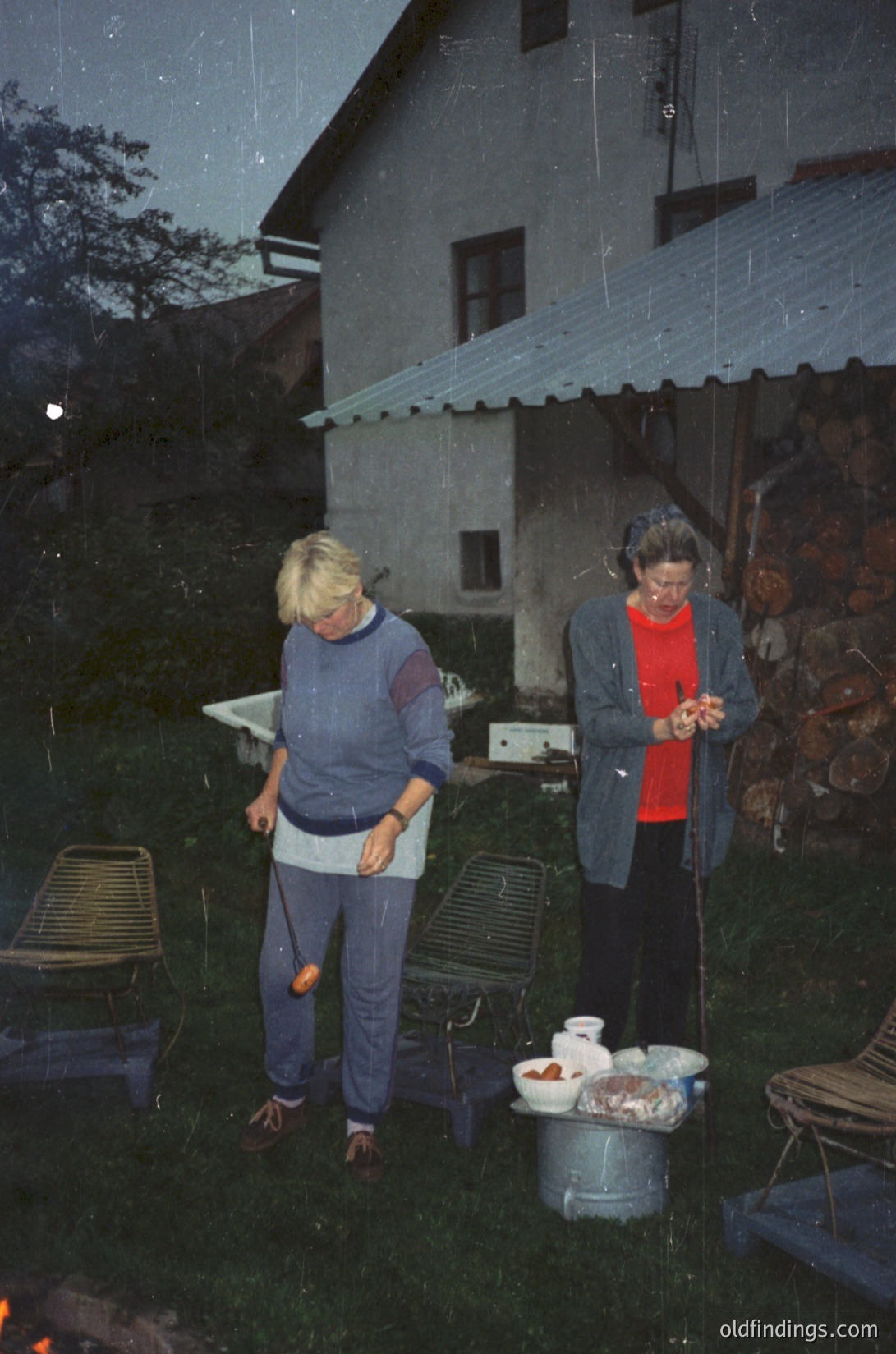 Two women in 1970s alpine retreat, preparing food under corrugated roof shelter. Left: woman in striped sweater and jeans holding orange object; right: woman in red jacket and headphones organizing ingredients in metal bucket. Foldable chairs and outdoor kitchen setup visible.