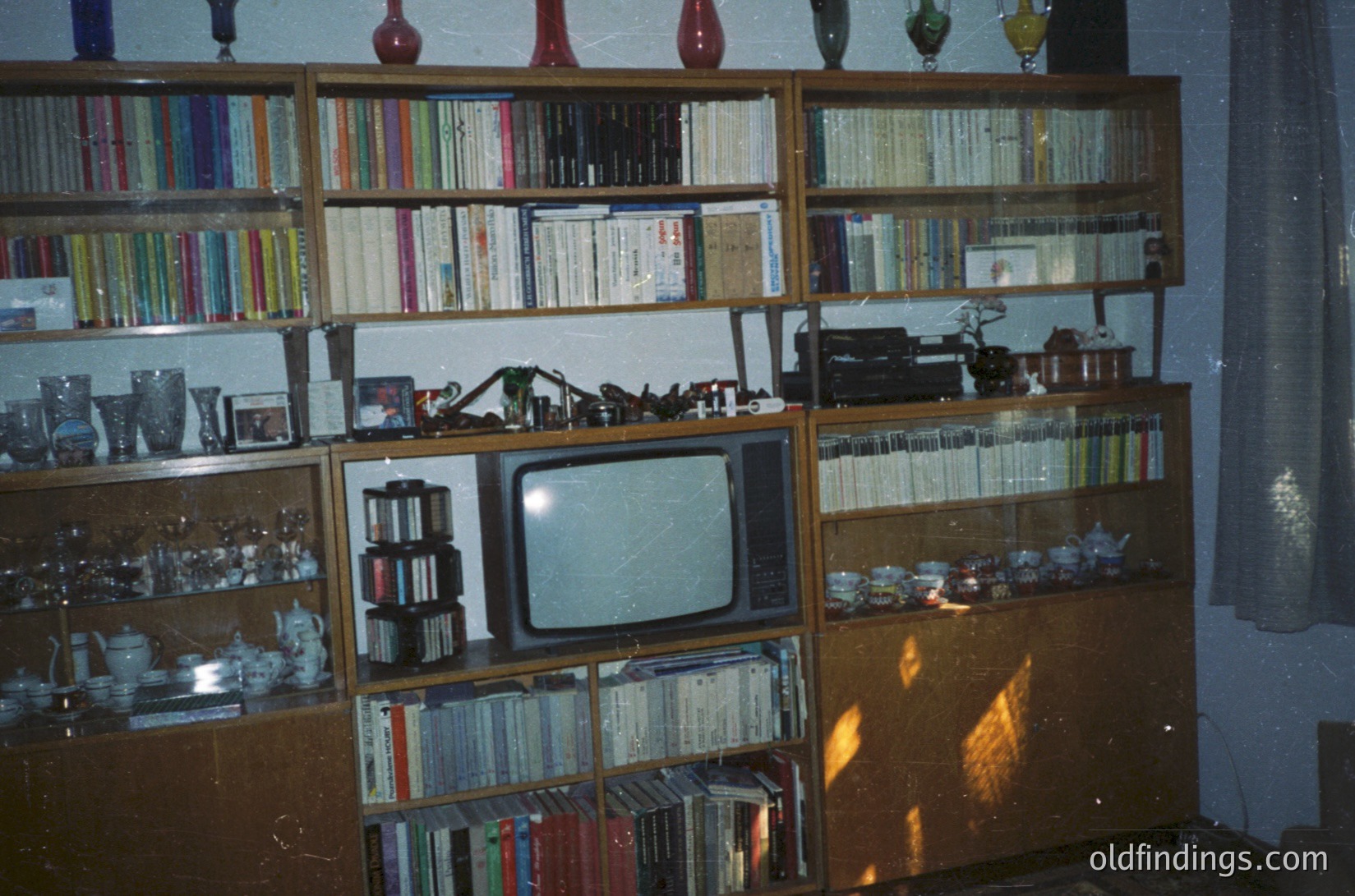 Vintage wooden bookshelf displaying mid-20th century domestic life. Shelves packed with books, vinyl records, and decorative glassware. Centerpiece: CRT TV with static, flanked by vintage radios and figurines. Warm lighting suggests evening or indoor photography. Likely Eastern European home, 1960s-1980s era.