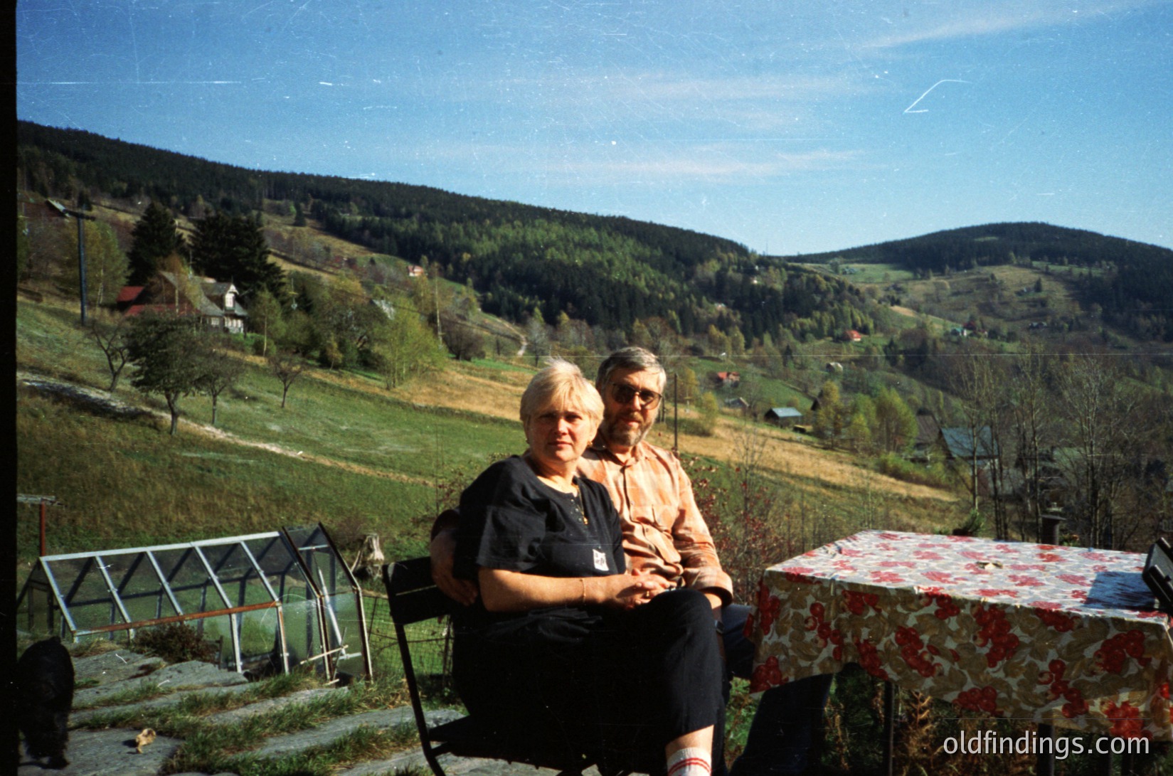 Two individuals pose outdoors on vintage wicker chairs beside a floral-patterned table, set against a lush, rolling countryside with scattered farmhouses and dense forests. The scene suggests a rural European setting, likely mid-20th century (1950s–1970s). [Vintage rural portrait with lush green valley and farmhouses, mid-20th century ]