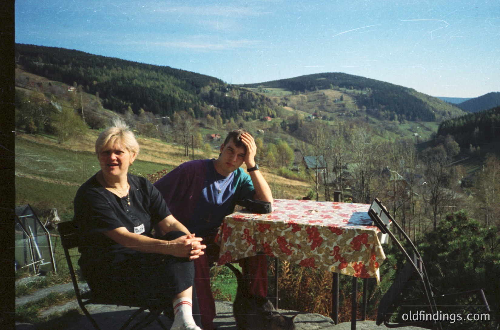 Two individuals pose outdoors at a rustic wooden table draped in floral-patterned red/white tablecloth, set on a concrete terrace. Lush green hills and forested slopes frame the background under clear skies. The scene suggests a rural European setting, likely 1990s–2000s.