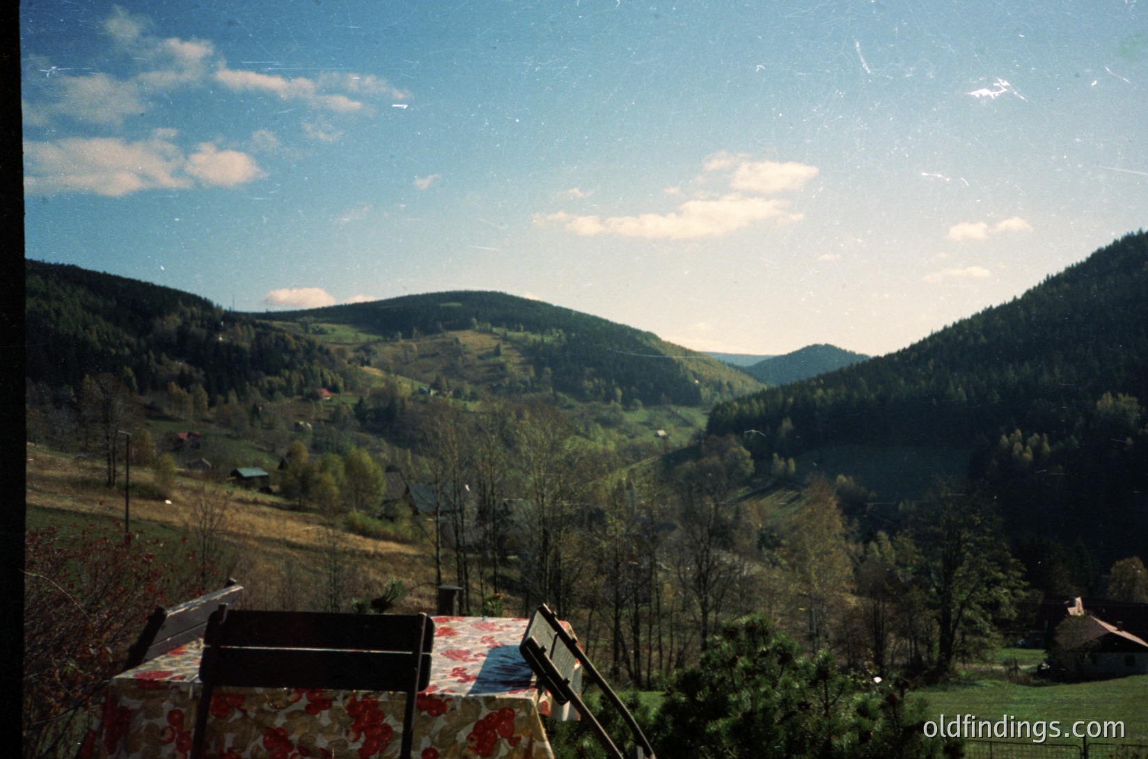 Vintage landscape shot of a valley with rolling hills, sparse forest, and scattered farmhouses. Foreground features a patterned picnic blanket and metal grill, suggesting outdoor dining. Soft color tones and slight film grain indicate 1970s–1980s era. Ideal for nostalgic or nature-themed content.
