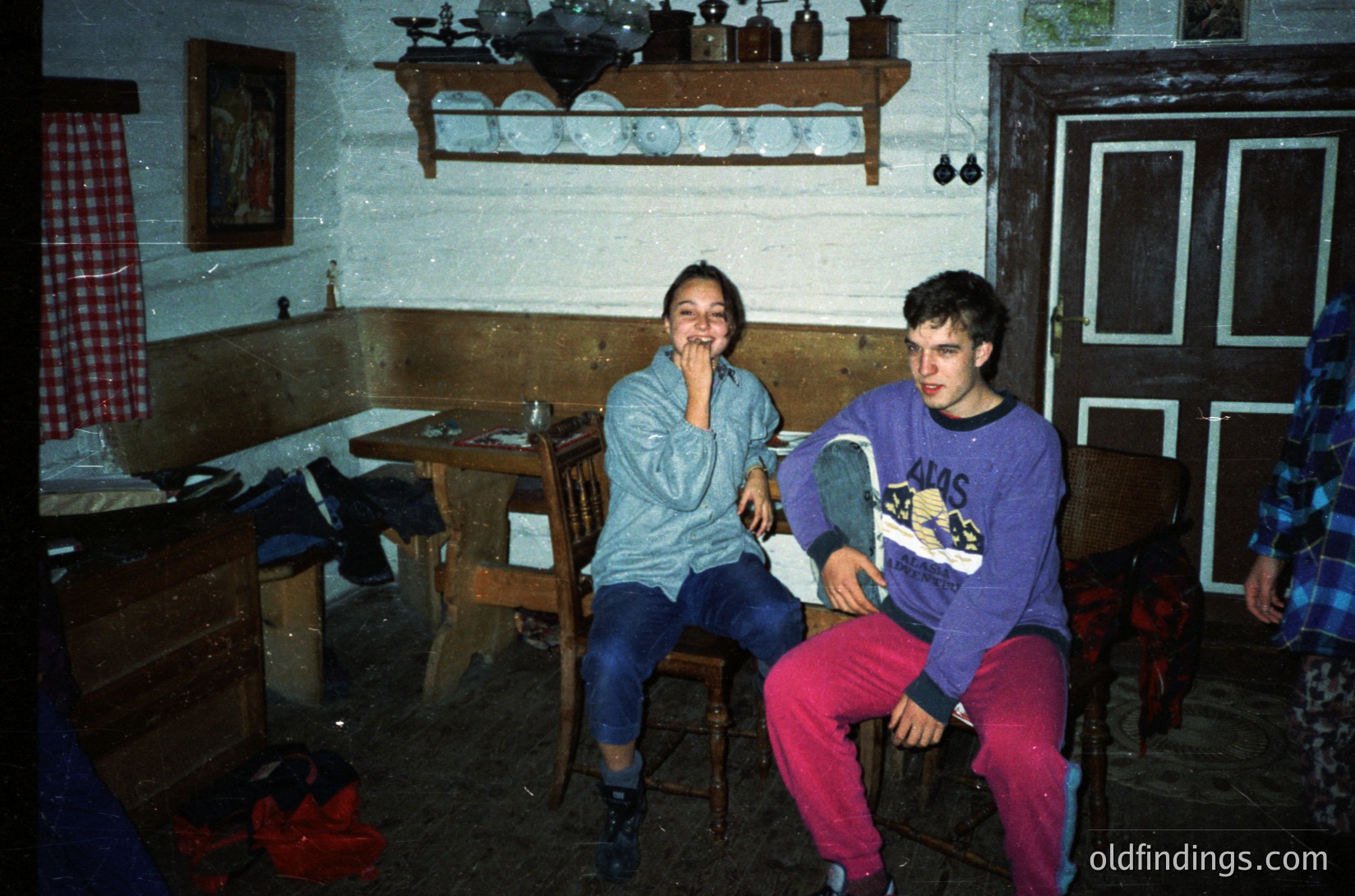 Vintage indoor scene featuring two individuals in a rustic, dimly lit room with wooden furniture. The girl wears a light blue sweater and blue jeans, while the boy sports a purple sweatshirt with a graphic print and red pants. Wooden shelves display ceramic jars and a framed painting.