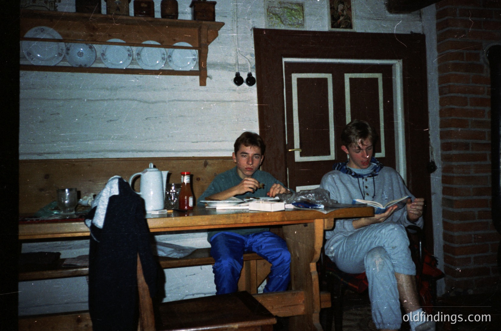 Vintage indoor study scene in rustic wooden cabin. Two young men seated at a wooden table with papers, pens, and a teapot; shelves display ceramic plates. Warm lighting and brick fireplace suggest a cozy, rural setting, likely Eastern Europe, 1970s–1980s.