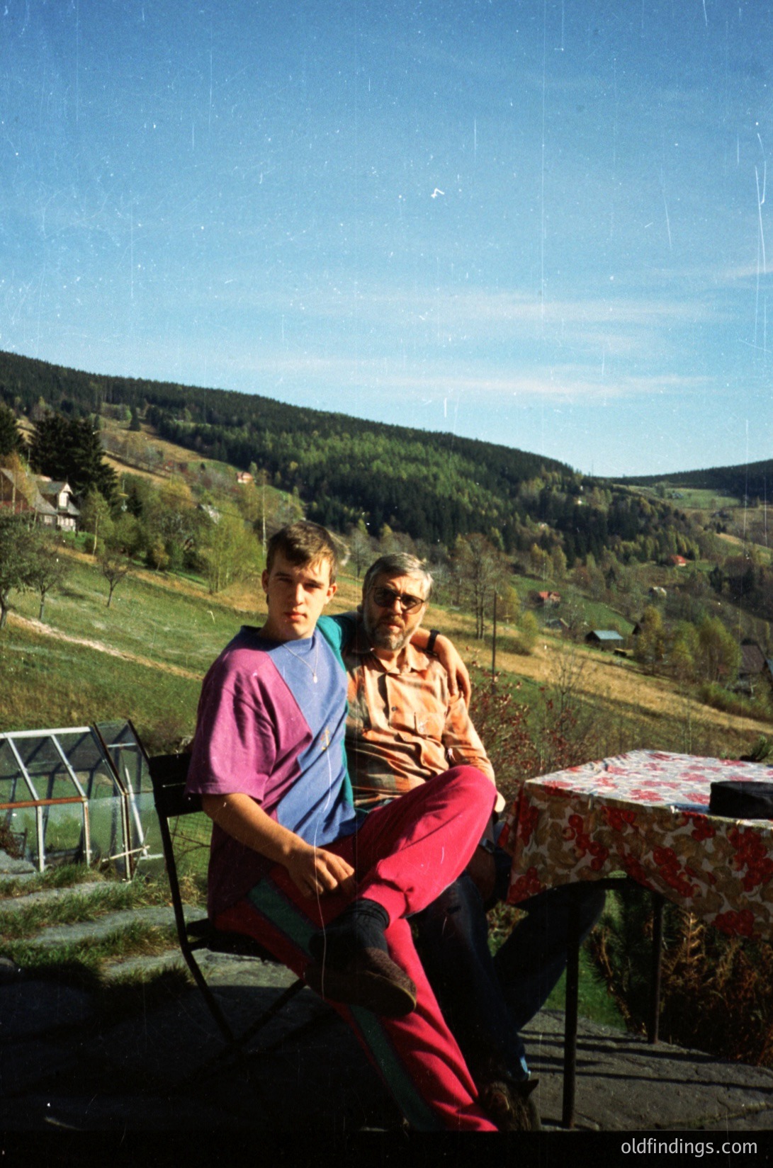 Vintage outdoor portrait of two individuals seated on wrought-iron chairs in a rural setting, likely Eastern Europe, 1970s. Man in striped shirt and red pants poses beside an elderly woman in patterned dress. Table with floral tablecloth and blurred greenery/road in background.
