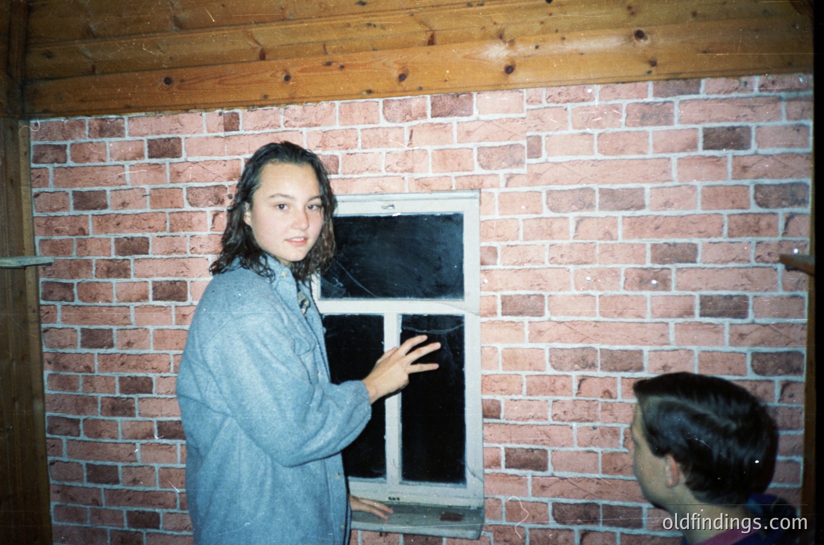 Young woman in a vintage denim jacket stands beside a small, framed window in a brick-walled room, framed by rough-hewn wooden beams above. Her pose suggests candid interaction with an unseen subject outside. The brickwork and window style suggest mid-20th-century residential architecture. [Vintage interior with brick wall and wooden beams, mid-20th century ]