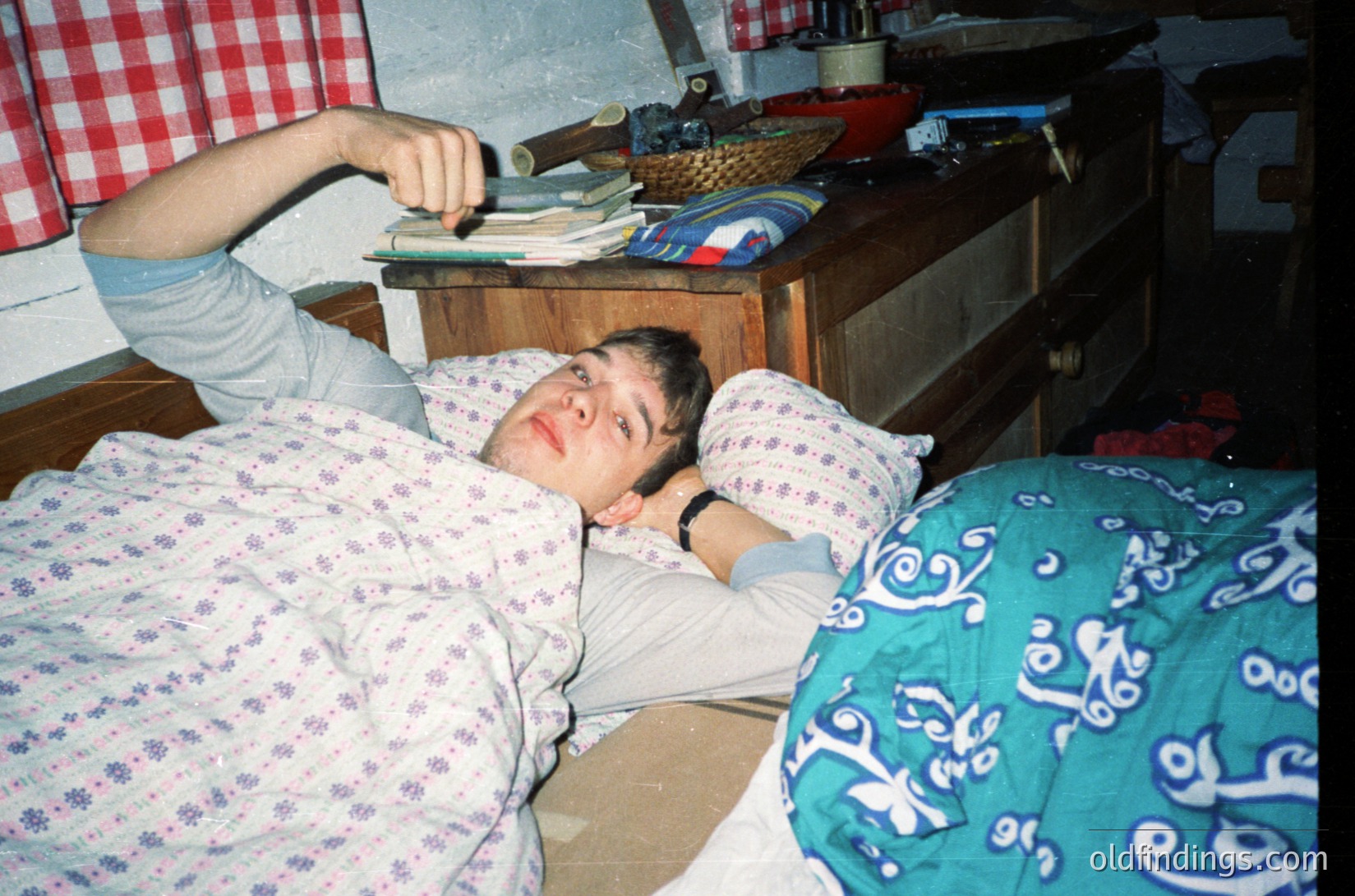 Vintage bedroom scene featuring a relaxed individual lying on floral-patterned bedding, holding a remote. Wooden furniture, including a nightstand with a basket and books, suggests mid-20th-century domestic decor. Checkered red/white tablecloth hints at retro home aesthetics.