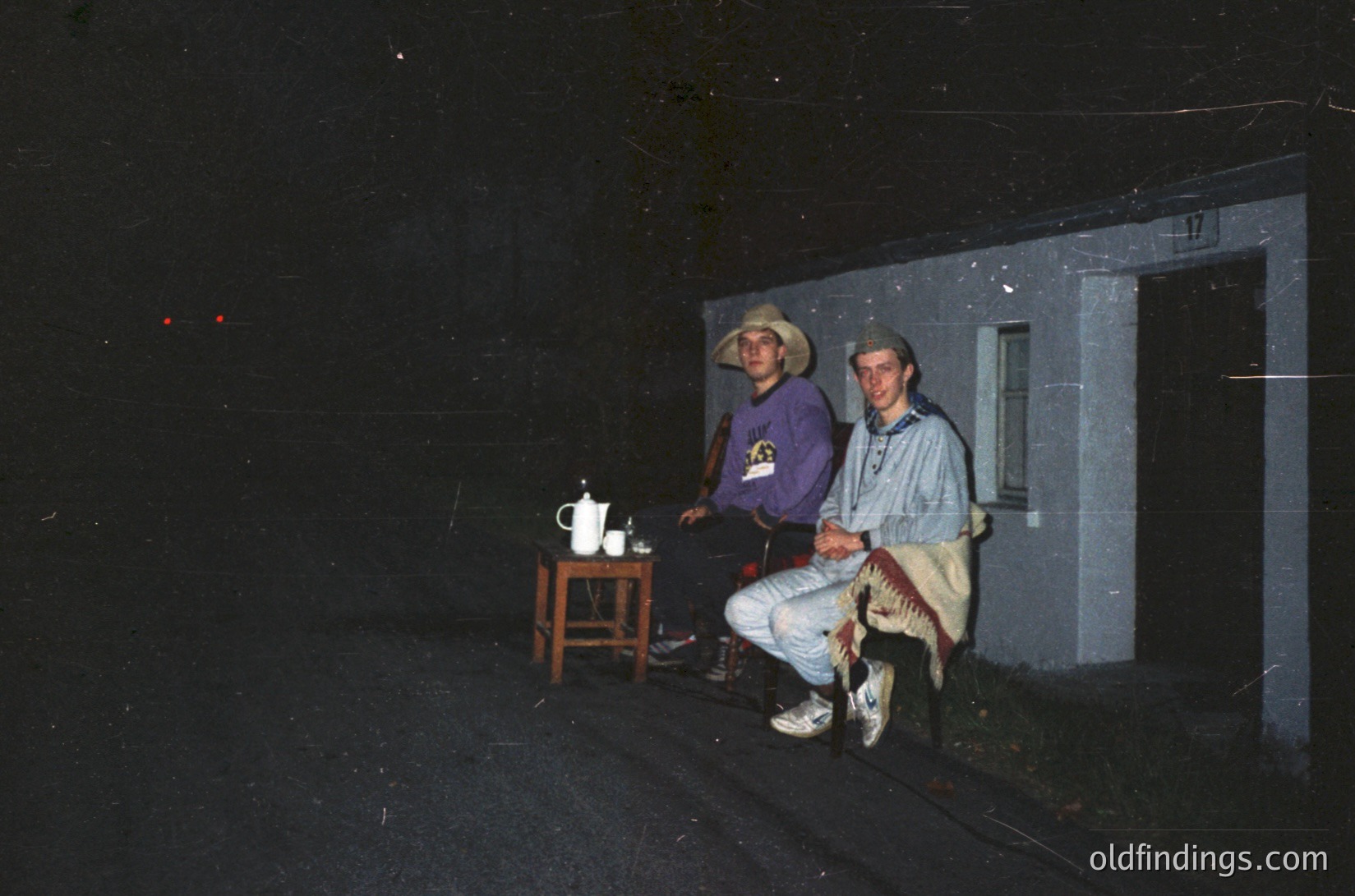 Vintage black-and-white photo of two men posing outside a plain concrete building. One wears a cowboy hat and a purple shirt with a logo, the other a beige jacket and light pants. A small wooden table with a teapot sits between them. Likely late 20th century, possibly 1970s–1980s.