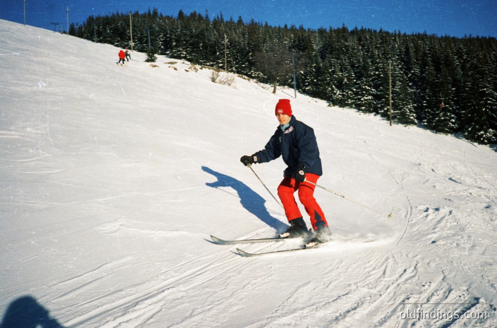 Vintage skier in bright red cap and matching ski pants descends groomed slope, mid-turn with poles raised. Forested alpine backdrop under clear skies. Likely mid-20th century European resort setting.